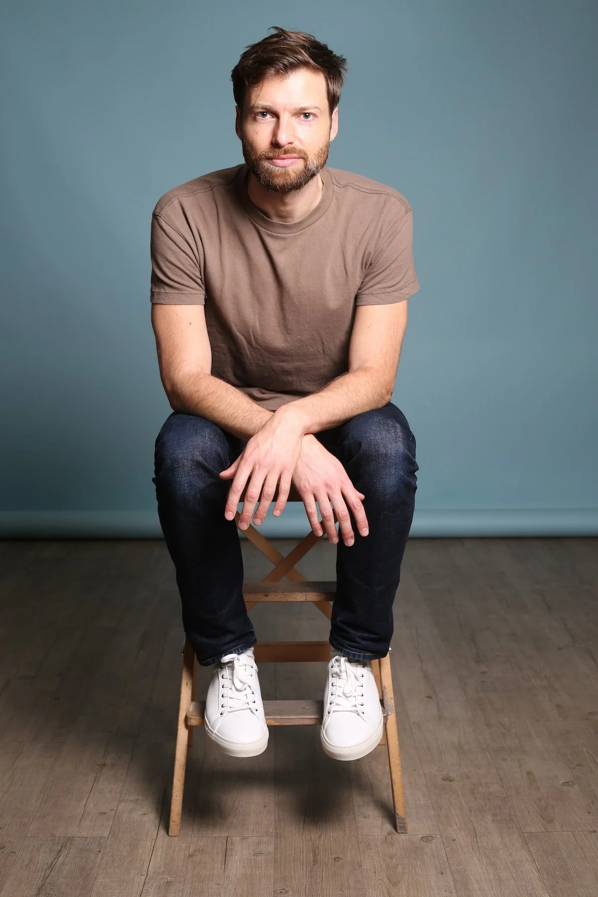 Stéphane Vernier assis sur une petite chaise en bois, portant un t-shirt beige, des jeans noirs et des baskets blanches, avec un fond bleu.