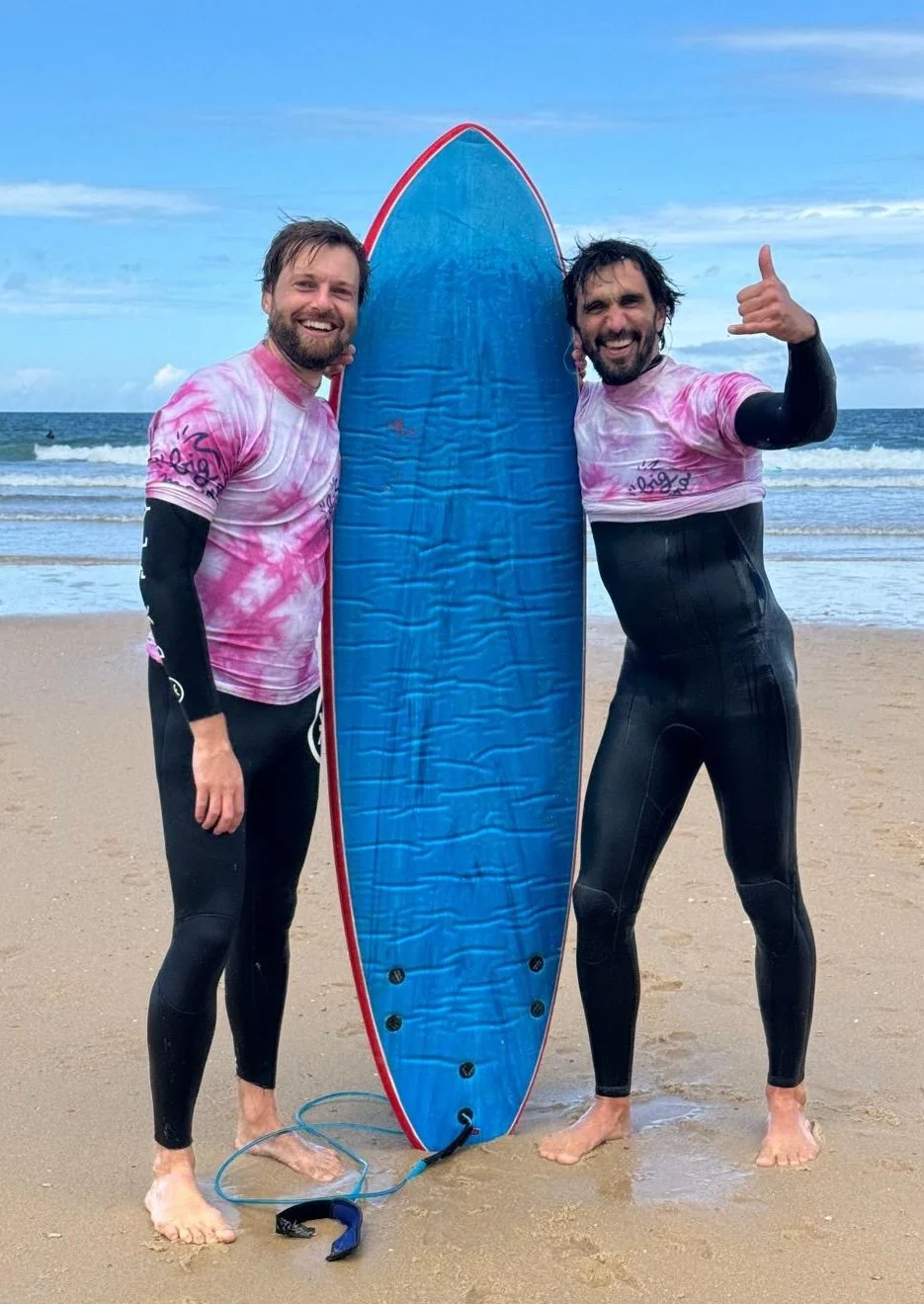Stéphane Vernier et Yoann Boyer en combinaisons de surf, souriants, posant avec une planche de surf sur la plage, sous un ciel bleu,  lors du stage La Sagesse des Vagues.