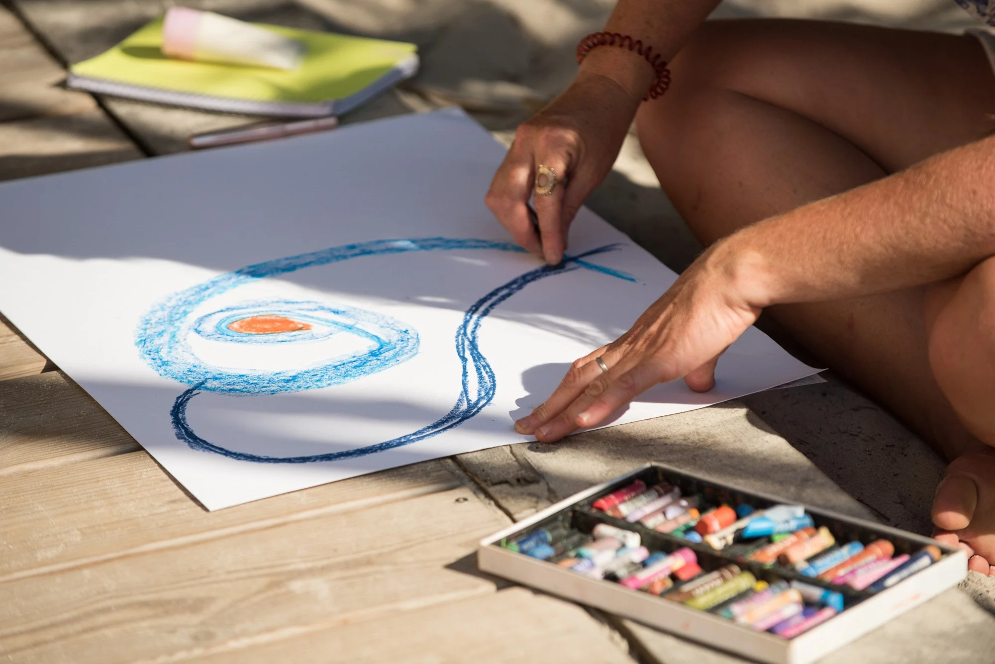 Personne en train de dessiner avec des pastels sur une feuille blanche sur un sol en pierre, avec un carnet et des pastels à proximité, lors du stage La Sagesse des Vagues.