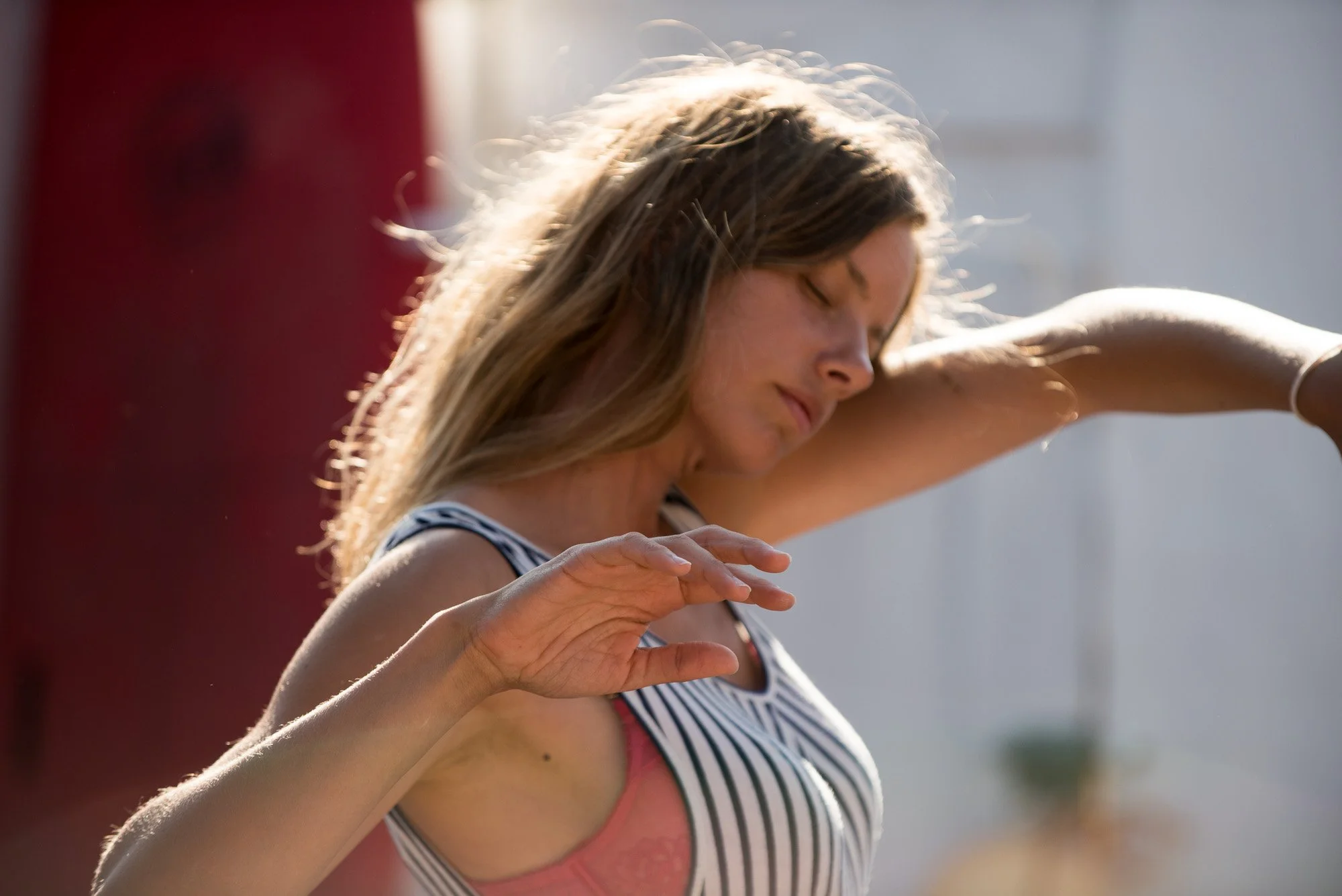Une femme avec les yeux fermés, portant un débardeur rayé, en plein mouvement avec le bras levé, sous une lumière naturelle en plein air.