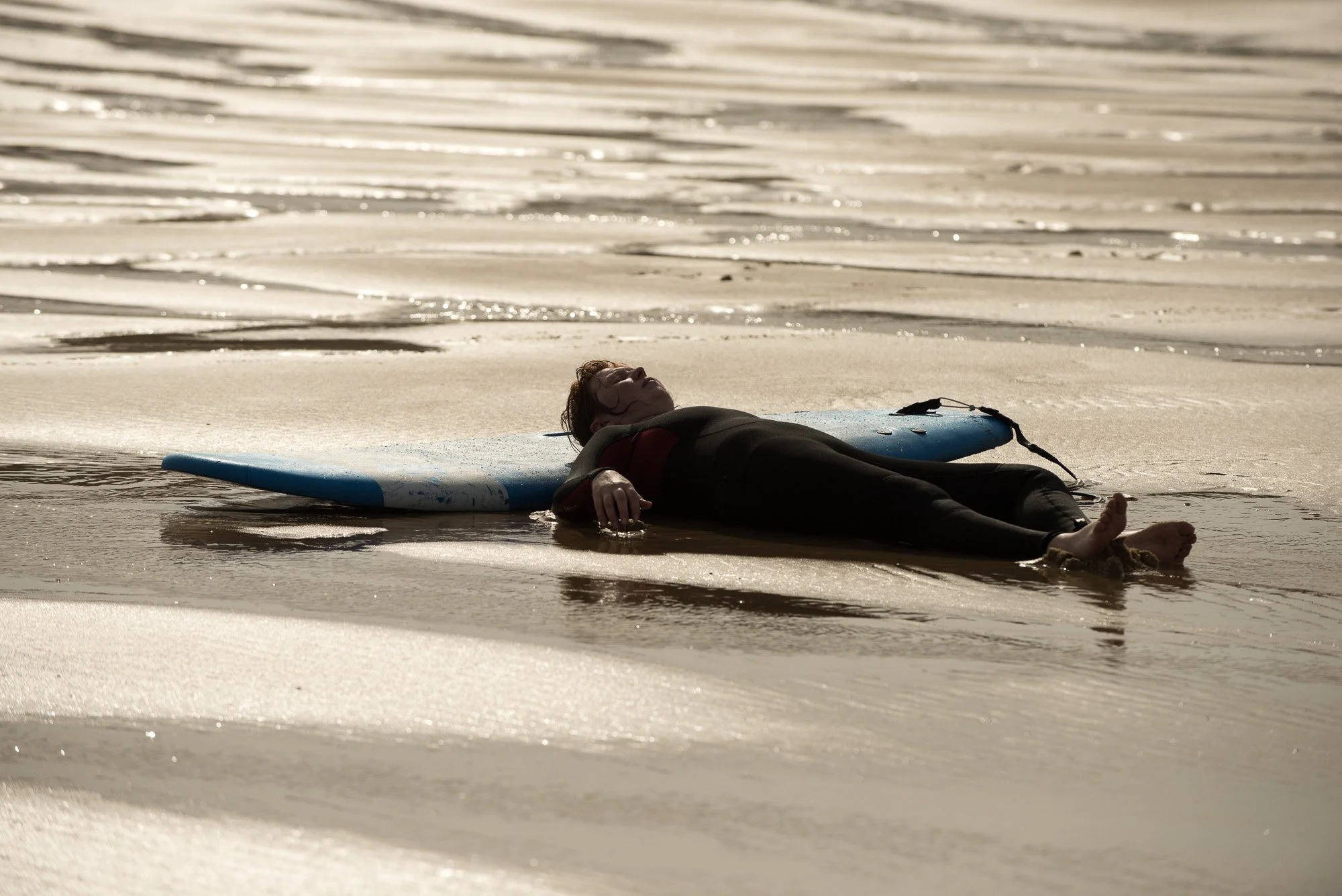 Une personne allongée sur sa planche de surf à plat sur le sable au bord des vaguelettes de l'océan.