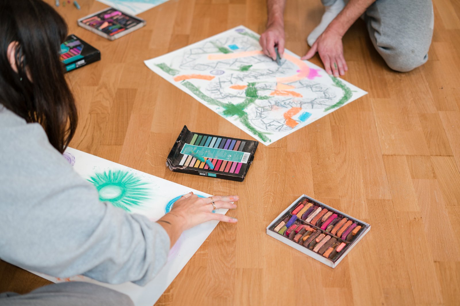 Deux personnes font de l'art avec des pastels et des crayons de cire sur des feuilles de papier, créant des dessins colorés sur un sol en bois, lors des dimanches Life Art Process® de Stéphane Vernier.