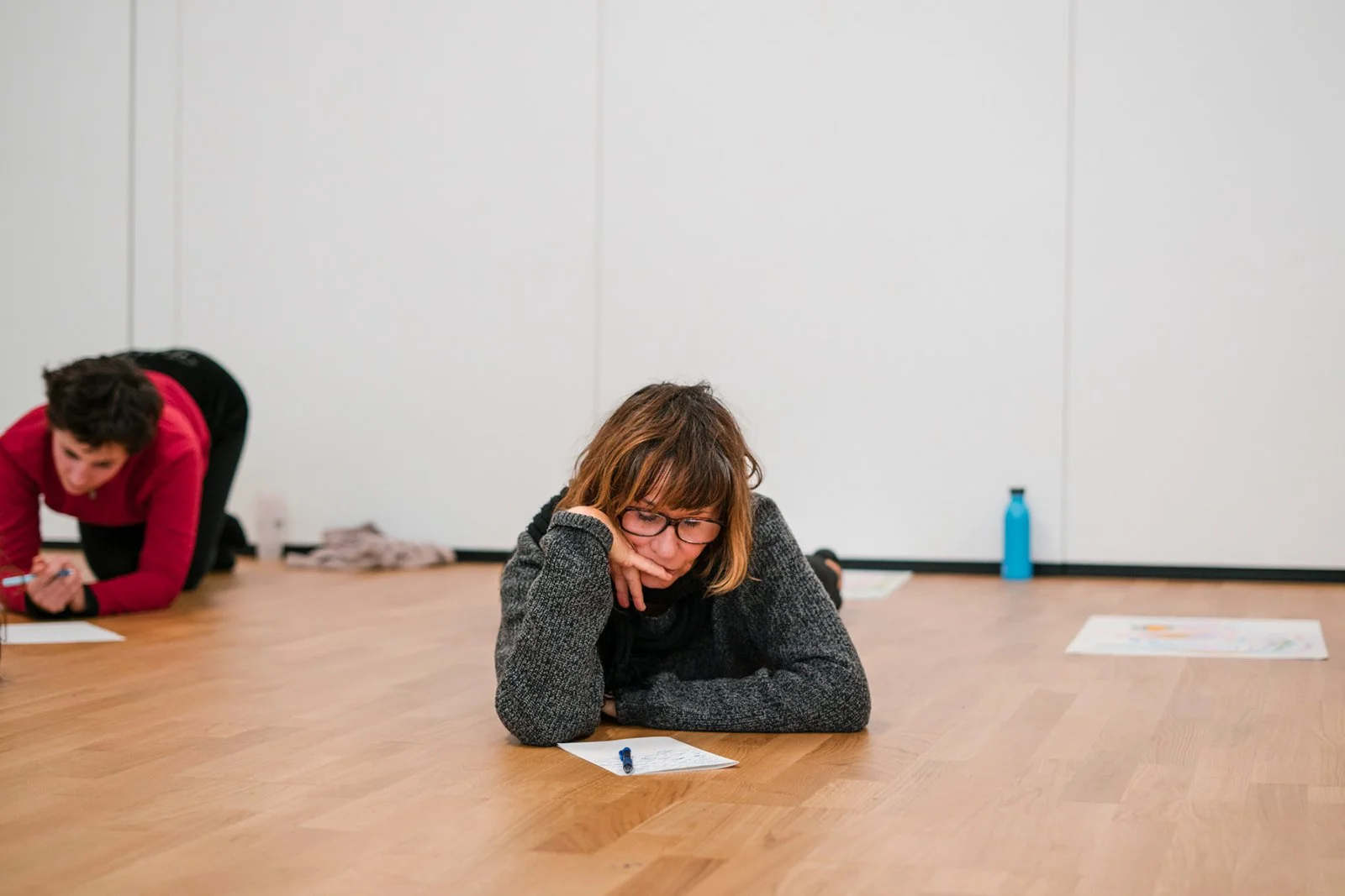 Deux personnes sont allongées sur un sol en bois, lisant ou travaillant sur des papiers avec stylos. La femme au centre porte des lunettes, une veste grise, et semble concentrée, tandis que l'autre personne à gauche porte un vêtement rouge et semble également concentrée.