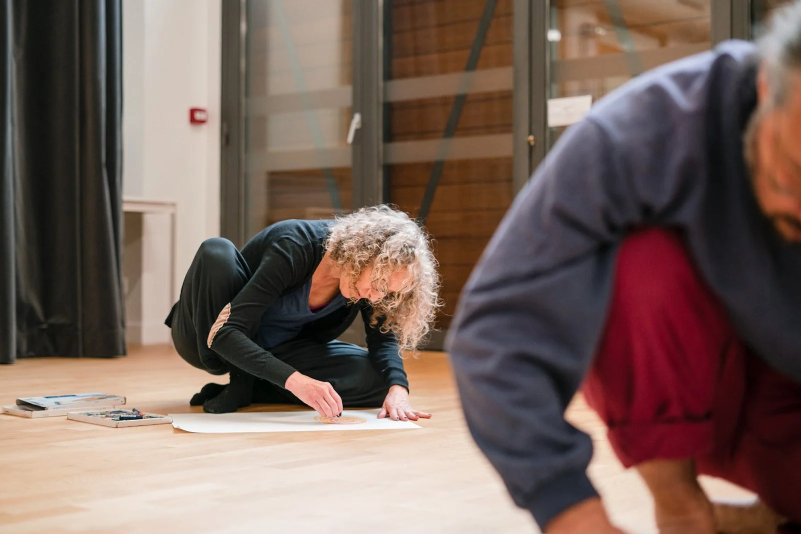 Deux personnes âgées, une femme avec des cheveux bouclés grisés et un homme avec des cheveux courts en arrière-plan, sont assises ou agenouillées sur un sol en bois, en train de créer une œuvre d'art avec des instruments de dessin.