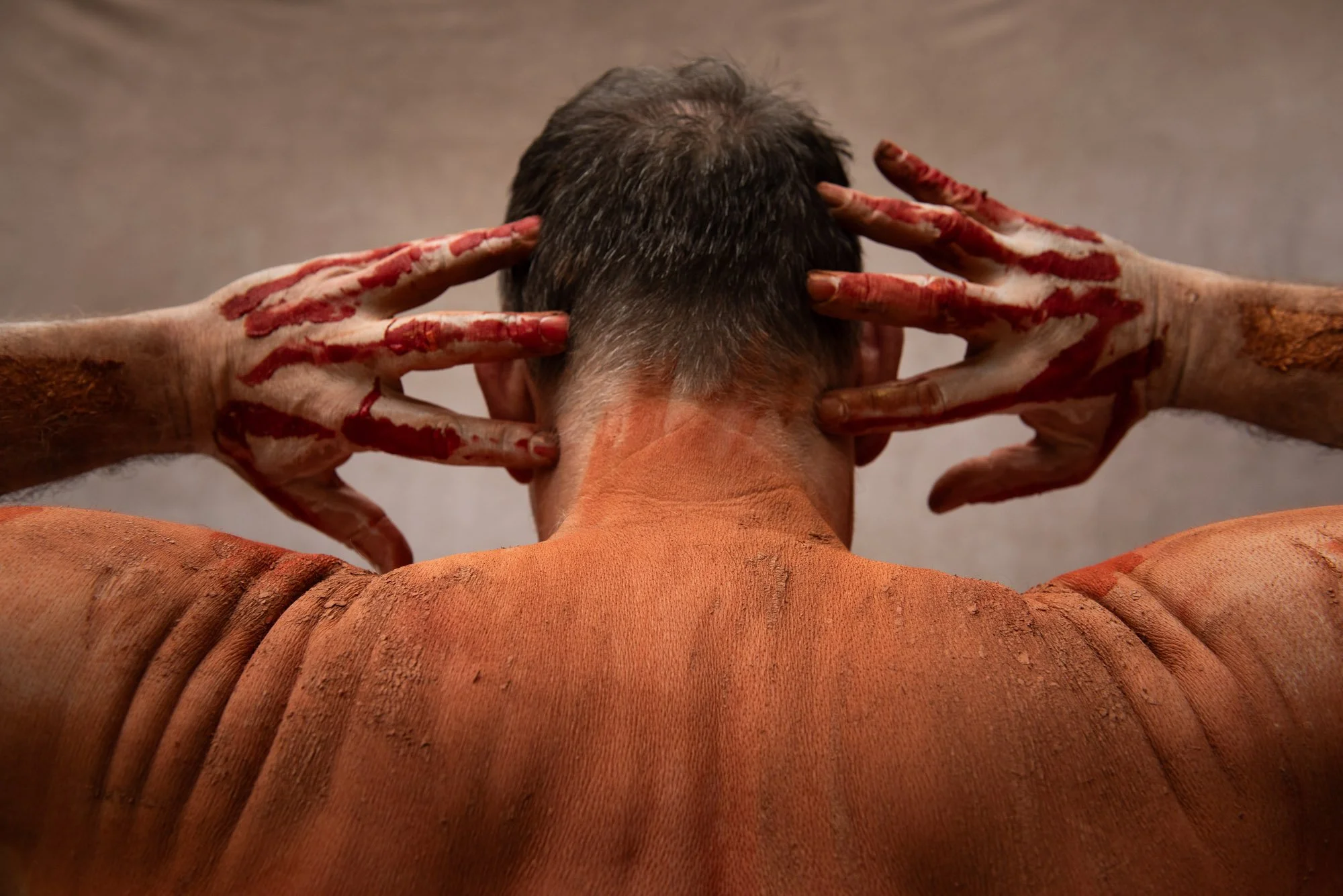 Une personne vue de dos, avec la tête inclinée en arrière, les mains couverte de peinture rouge, positionnées derrière la tête, dans un fond neutre, lors du stage Regards animé par Isabelle Chapuis et Stéphane Vernier.