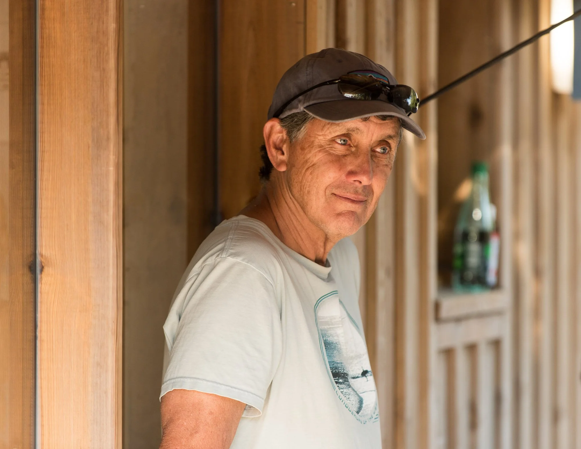Ken Otter porte un t-shirt blanc, une casquette et des lunettes de soleil, il se tient à côté d'un mur en bois, dans un cadre intérieur lumineux,  lors du stage La Sagesse des Vagues.