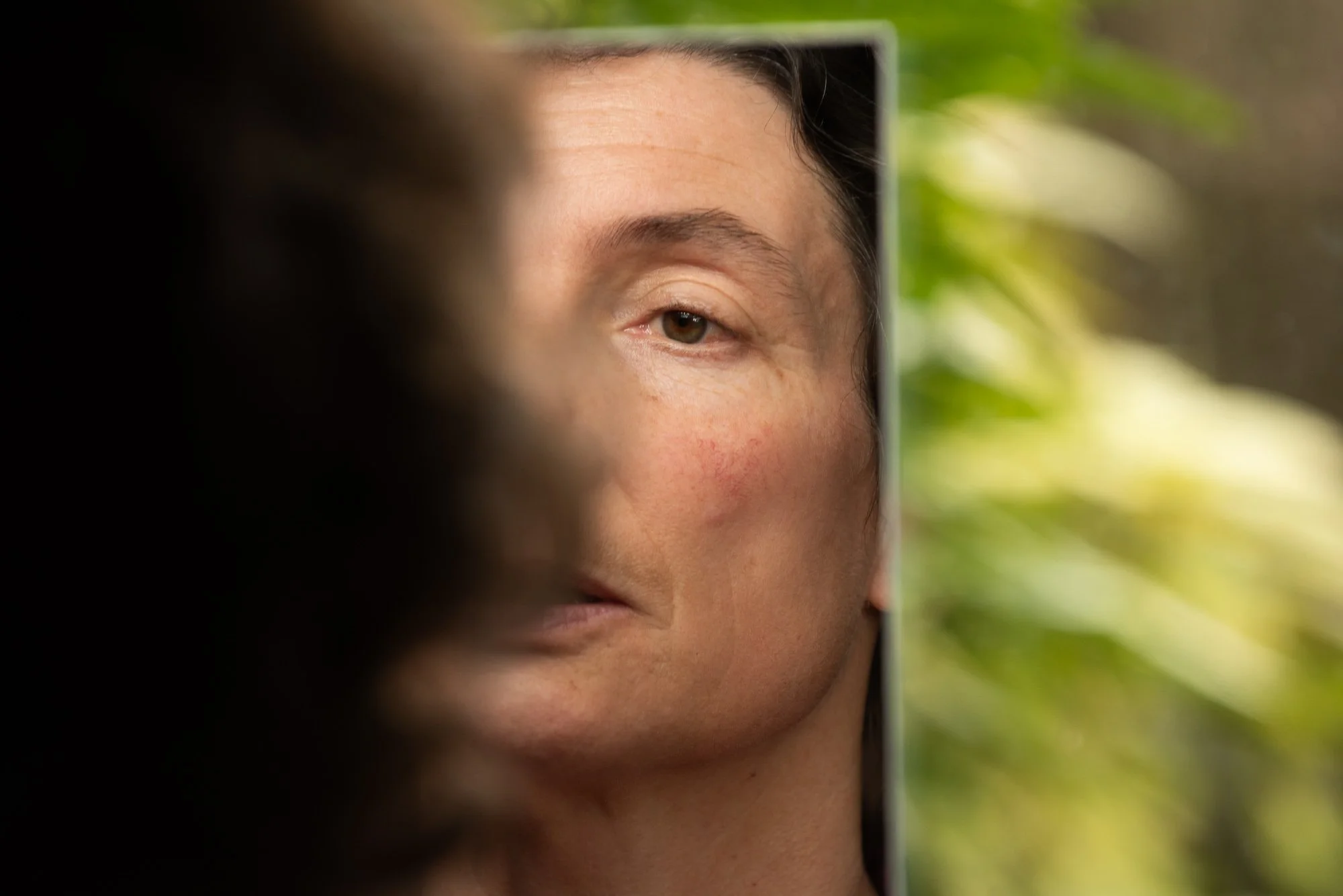Une personne regarde dans un miroir, révélant une seule moitié de son visage, entourée de végétation verte en arrière-plan, lors du stage Regards animé par Isabelle Chapuis et Stéphane Vernier.