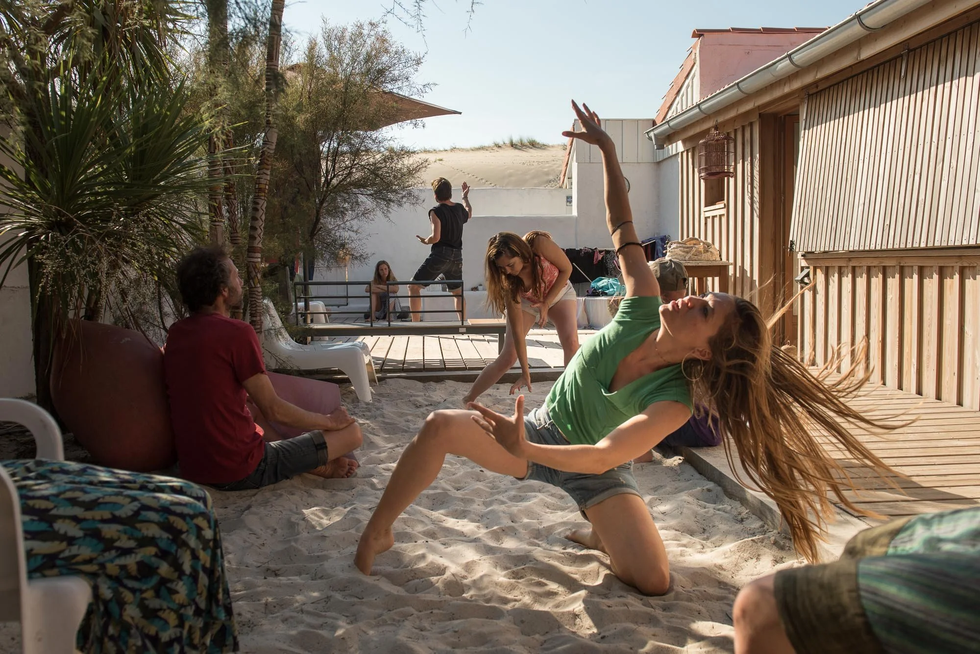 Un groupe de personnes en pleine exploration de danse, dans le sable, avec une maison en bois et un arbre en arrière-plan, lors du stage La Sagesse des Vagues.