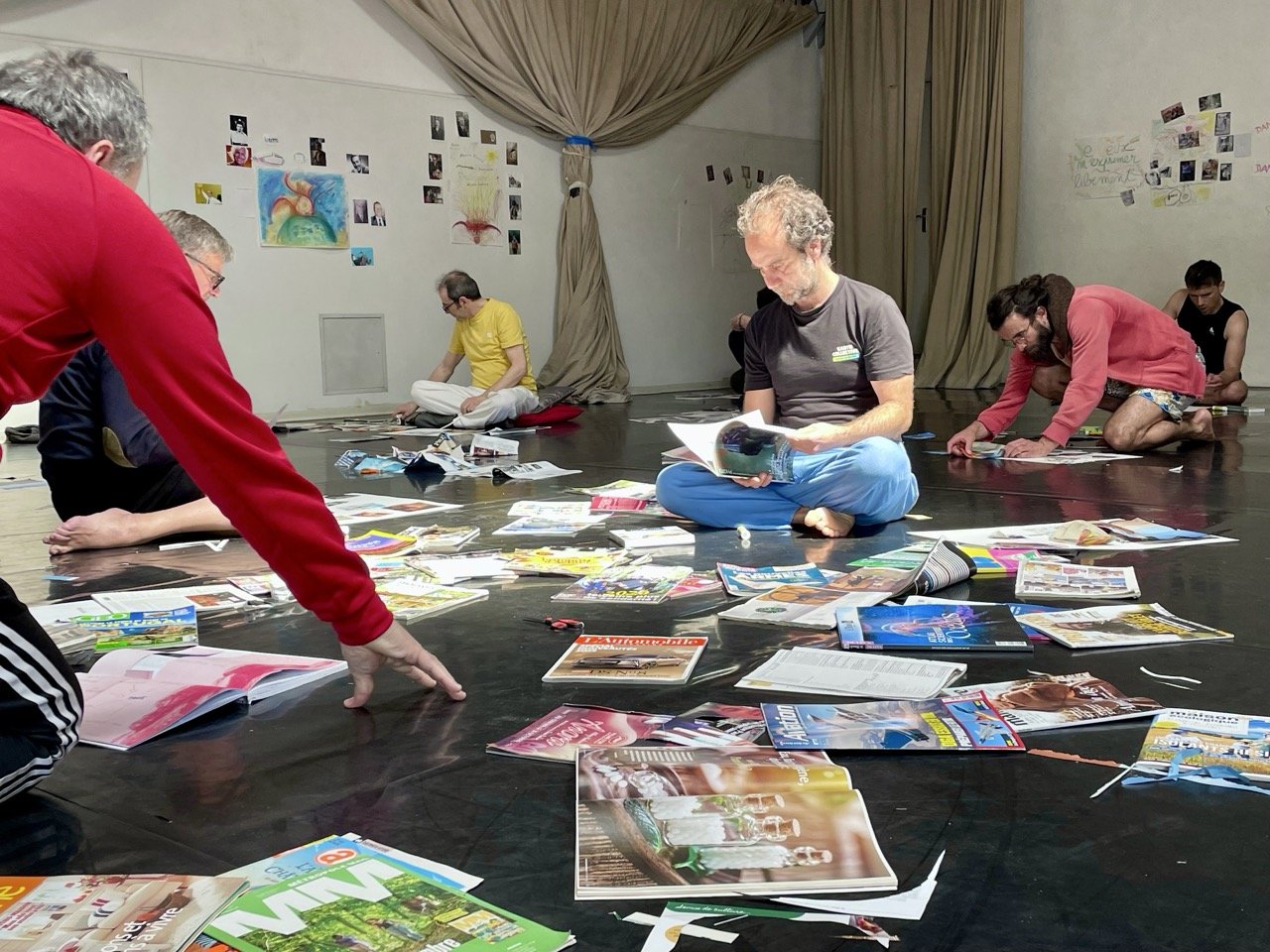 Groupe d'hommes assiss ou agenouillés sur un sol noir, entourées de magazines et d'articles, dans une salle avec des murs blancs décorés de dessins, photos et écritures, pendant le stage Life Art Process® Hommes.