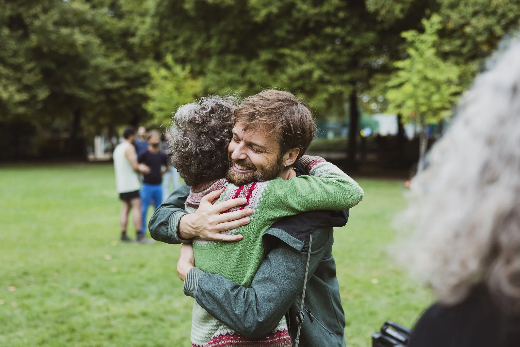 Stéphane Vernier embrasse une stagiaire dans un parc, avec d'autres personnes en arrière-plan, arbres verts et ciel.
