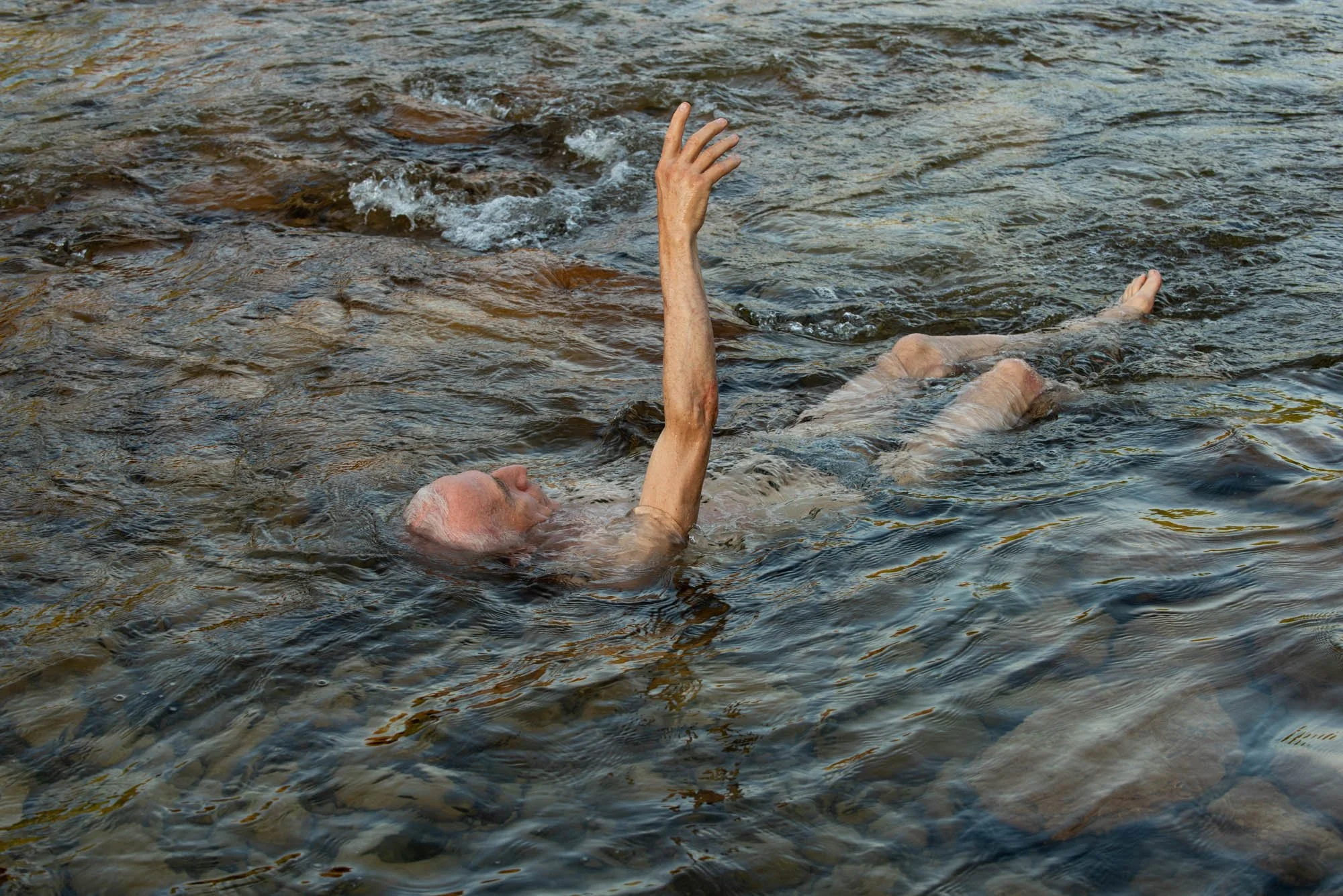 Une personne âgée nage dans une rivière avec un bras levé, l'eau est claire avec des pierres visibles au fond, lors du stage Nature.s avec Stéphane Vernier et Isabelle Chapuis.