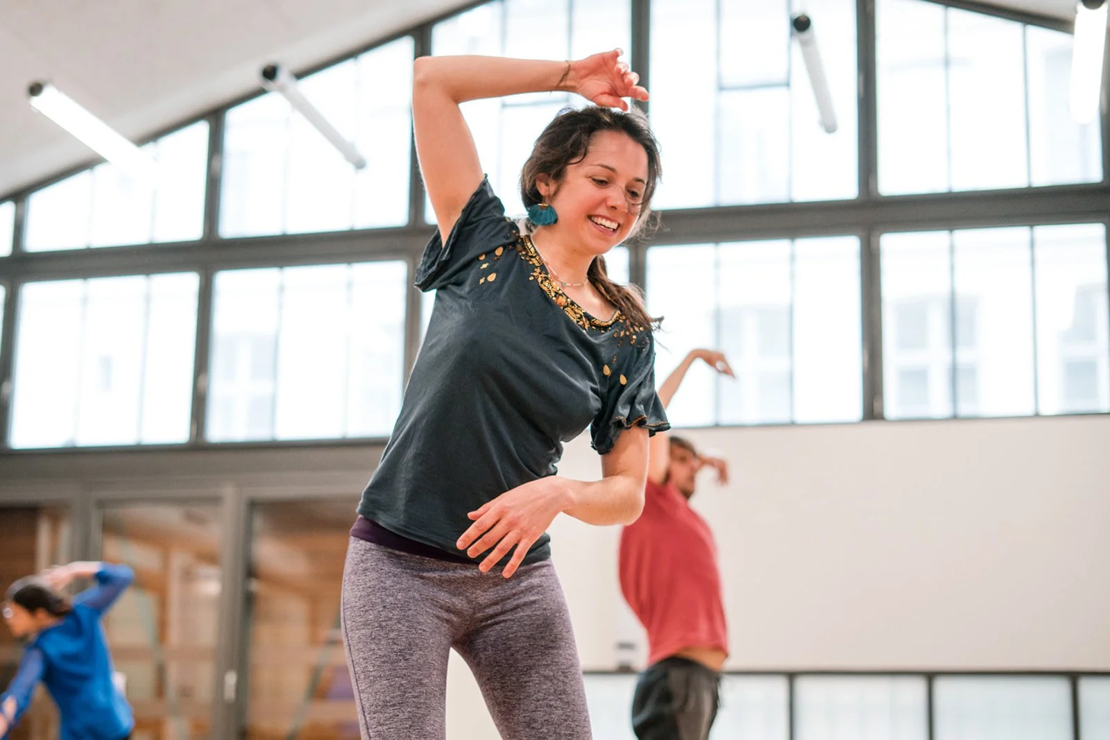 Une femme souriante en tenue de sport danse dans une salle lumineuse avec de grandes fenêtres, entourée de deux autres personnes également en mouvement.