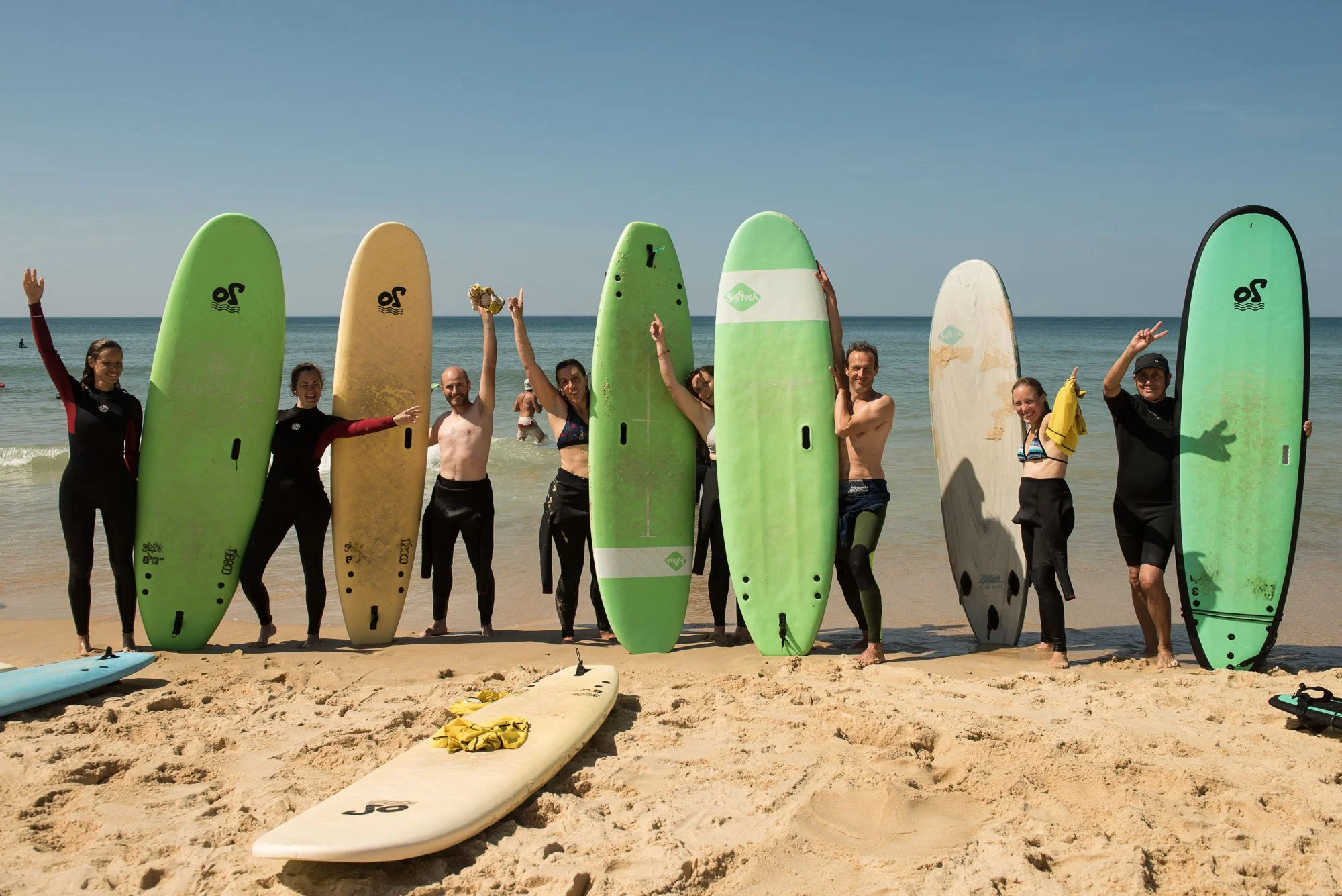 Groupe de personnes de différentes ethnies souriantes avec des planches de surf sur une plage de sable, onde océane en arrière-plan, sous un ciel clair, lors du stage La Sagesse des Vagues.