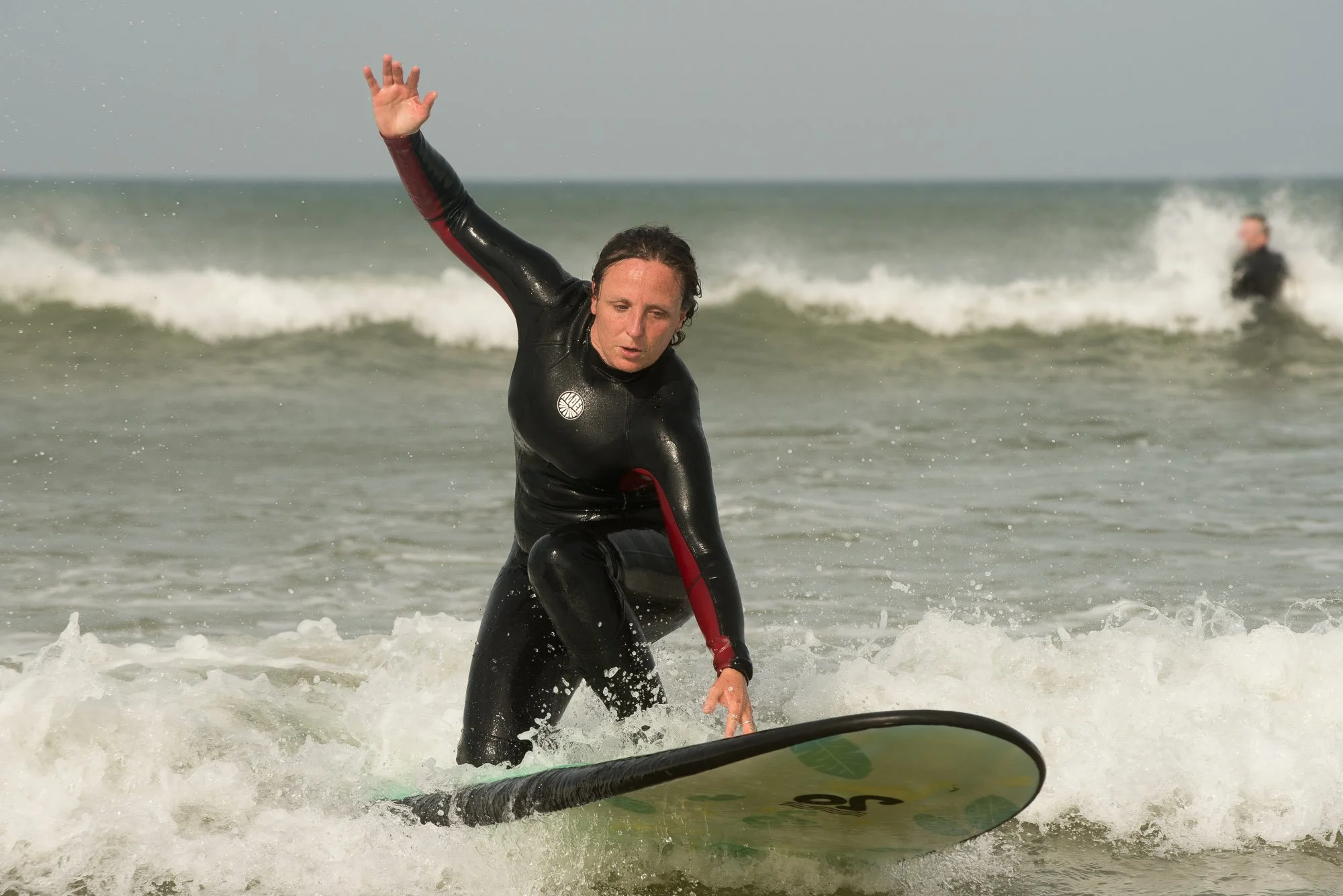 Femme en combinaison de surf, debout sur une planche, en train de surfer dans l'océan, levant une main en l'air, lors du stage La Sagesse des Vagues.