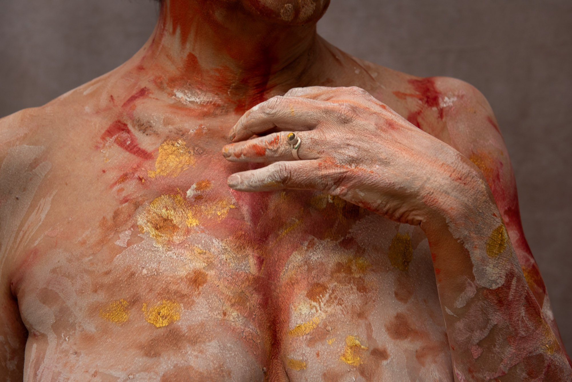 Une personne nue dont la peau est peinte avec des touches de peinture rouge, blanche et dorée, tenant sa poitrine avec une main, lors du stage Regards animé par Isabelle Chapuis et Stéphane Vernier.