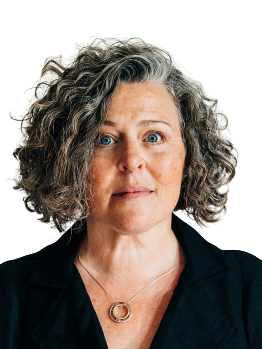 Close-up of a middle-aged woman with curly grey hair, blue eyes, wearing a black shirt and a silver circular pendant necklace, looking directly at the camera.