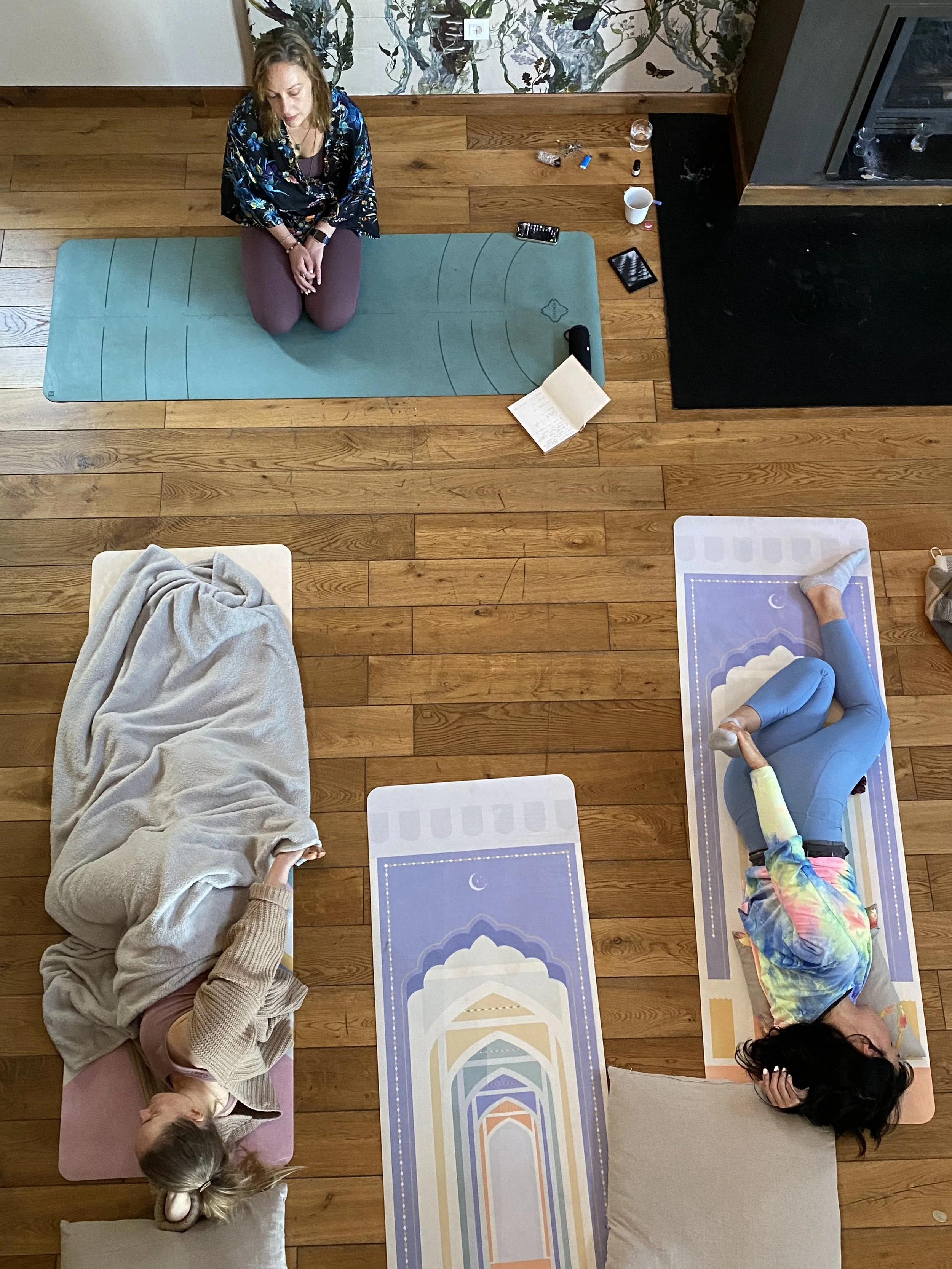 Three women in a yoga session, one sitting on a yoga mat, the other two lying on their sides on mats. The woman sitting appears to be guiding the session, sitting on a teal mat, while the other two are on patterned mats with a pastel motif. Various i