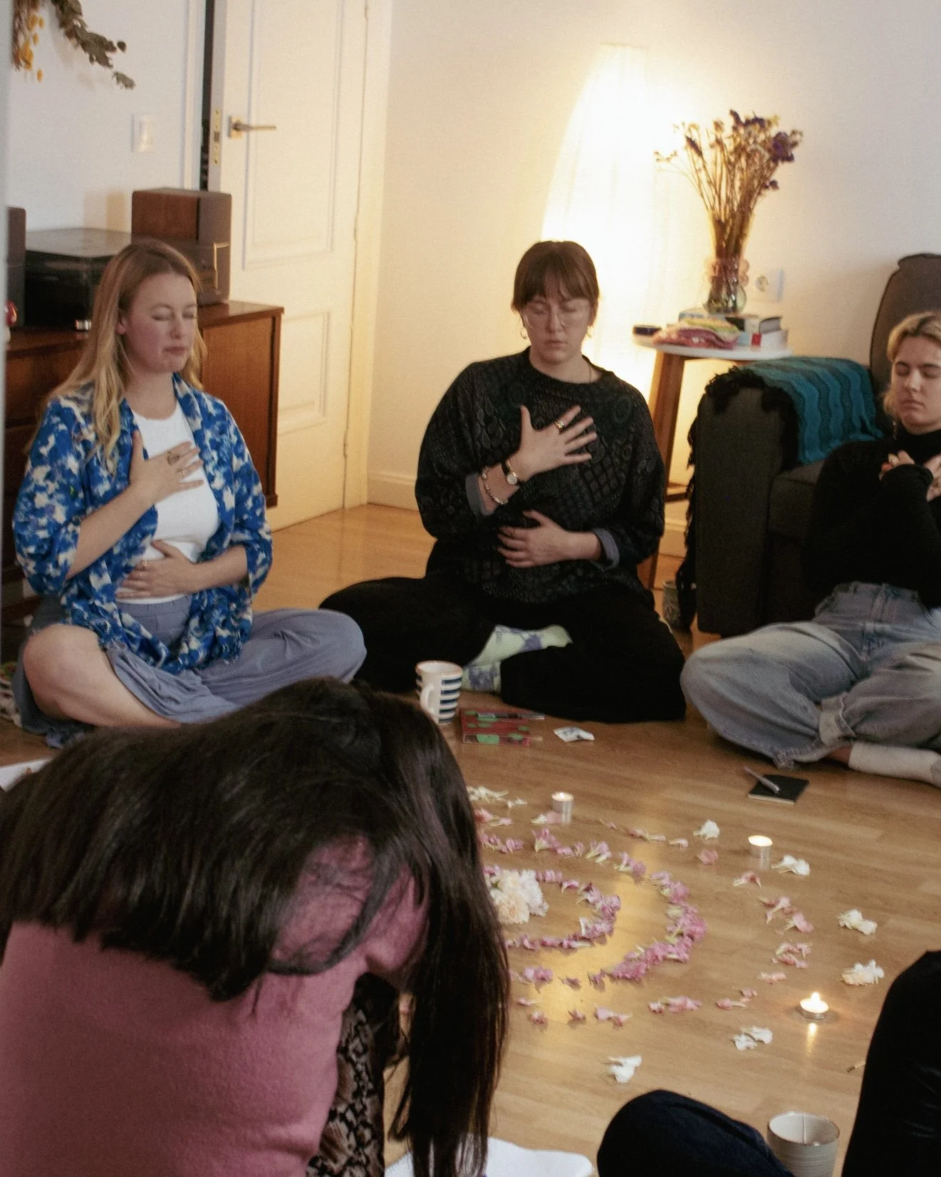 A group of women sitting cross-legged on a wooden floor in a circle, with candles and pink flower petals arranged in a spiral pattern, engaging in a meditation or mindfulness activity in a cozy living room.