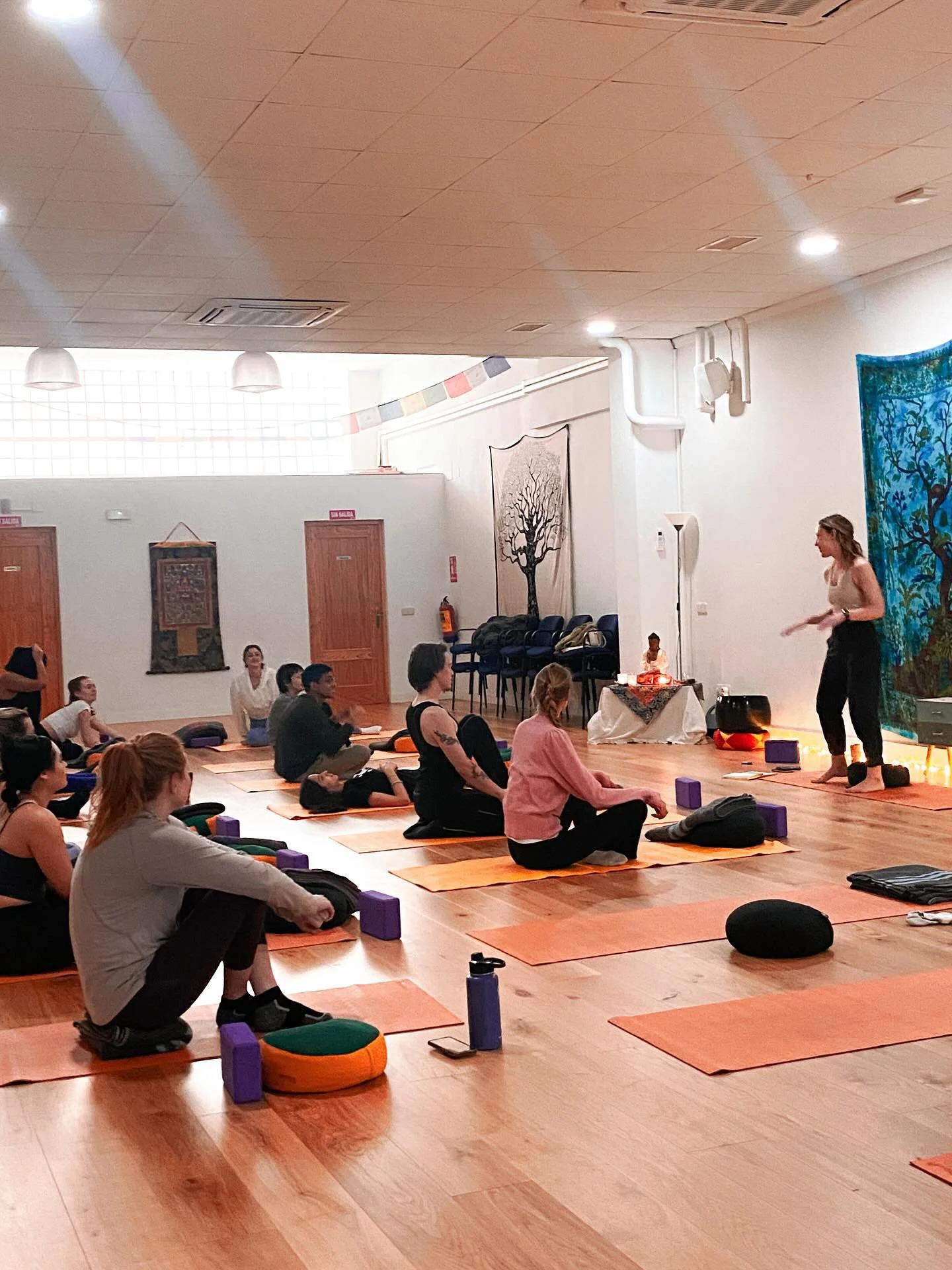 A yoga class in a spacious room with wooden floors. Participants are sitting on orange yoga mats, facing a female instructor near the front of the room. Some participants have yoga blocks, water bottles, and cushions nearby. The room features artisti