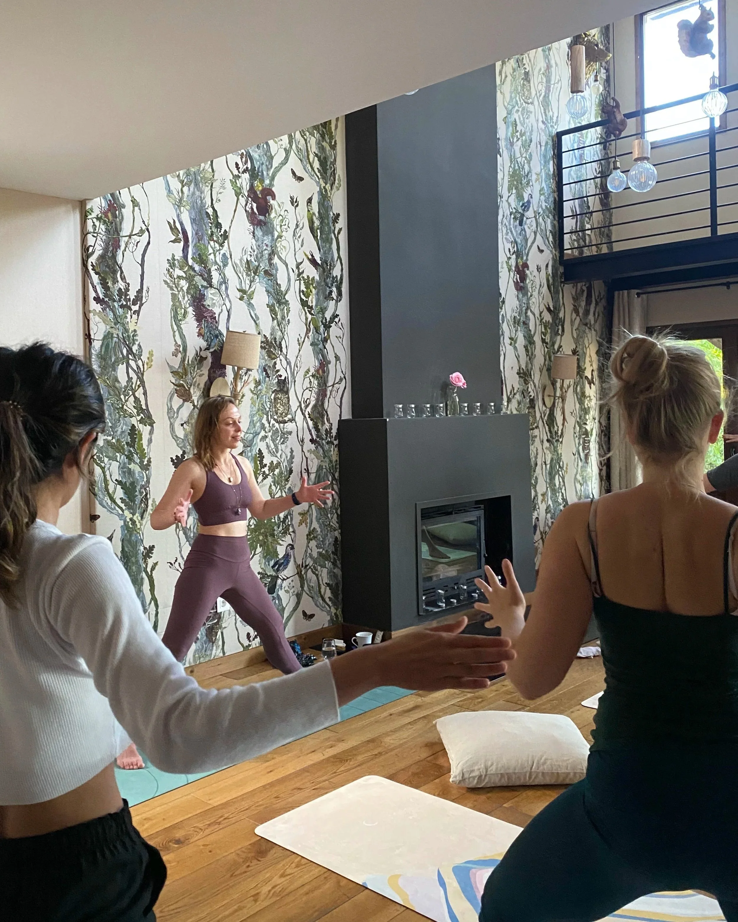 A woman leading a yoga class in a living room with floral wallpaper, a modern fireplace, and a high ceiling. Several participants are following her pose on yoga mats.