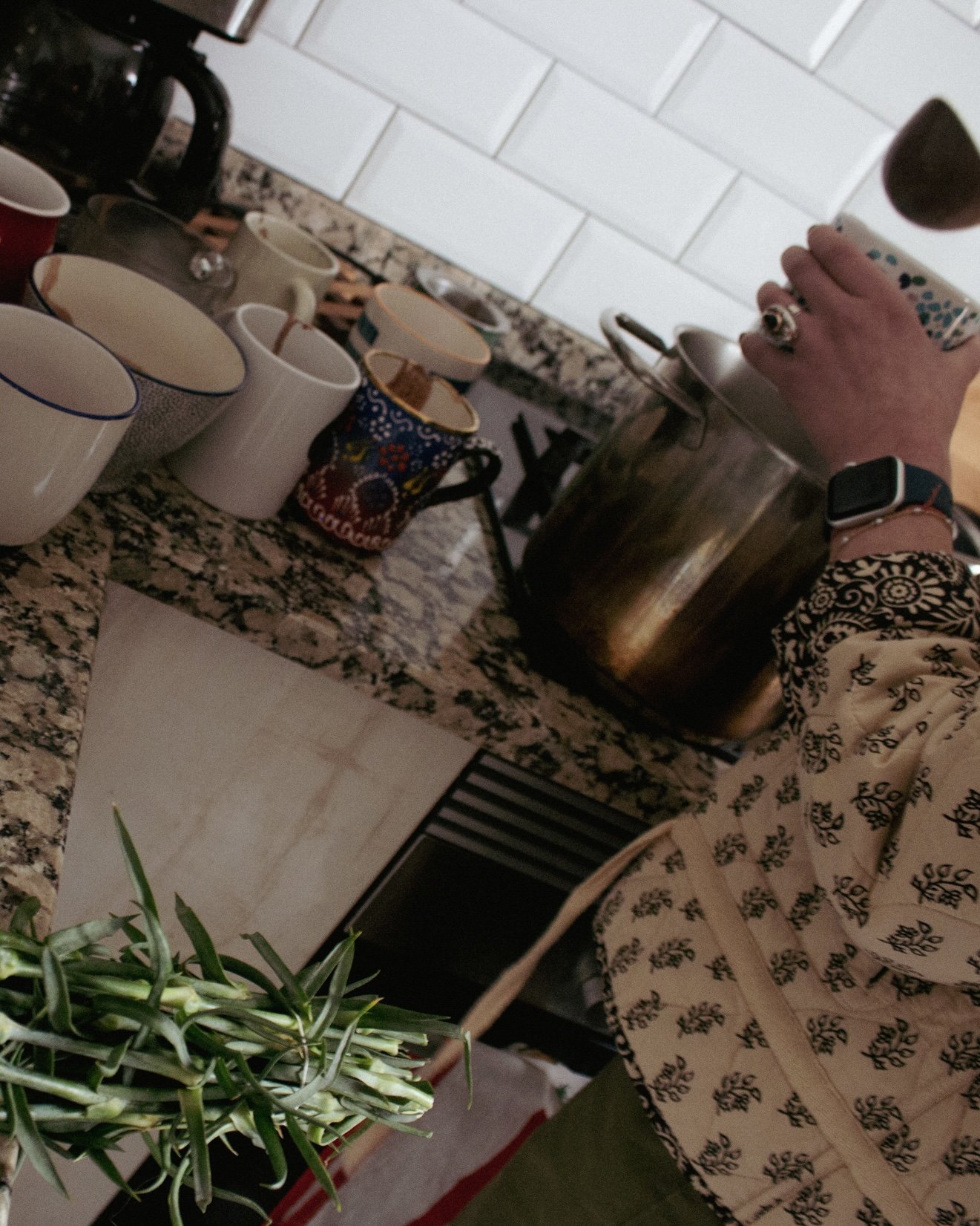 A person pouring ceremonial cacao into a mug from a large metal kettle on a kitchen countertop with several empty mugs and teacups nearby.