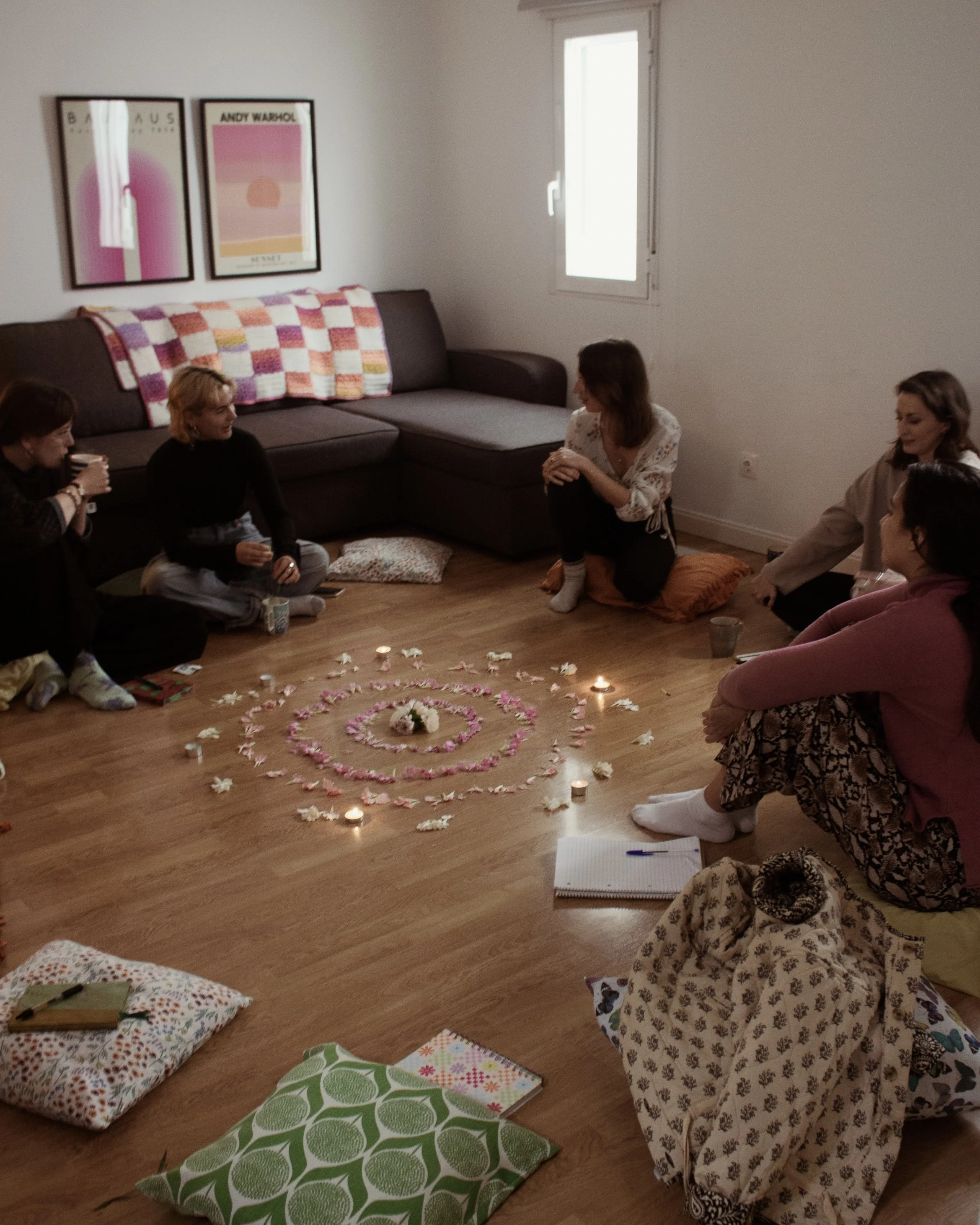 A group of people sitting in a circle on the floor around a decorative arrangement of flowers, candles, and petals, in a cozy living room with a sofa and wall art.