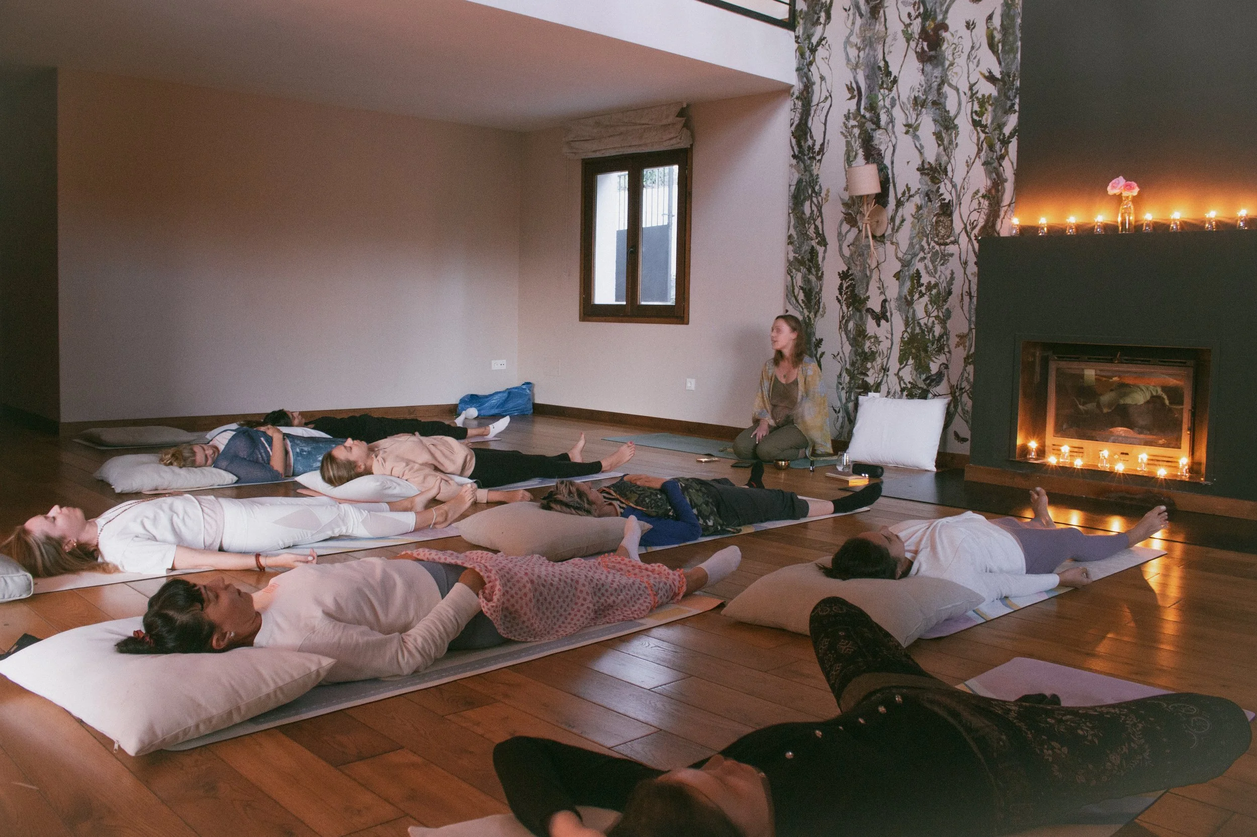A group of people participating in a yoga or meditation class in a cozy room with hardwood floors, a fireplace, and decorative wallpaper, lying on mats with pillows, guided by an instructor sitting on the floor near a pillow.