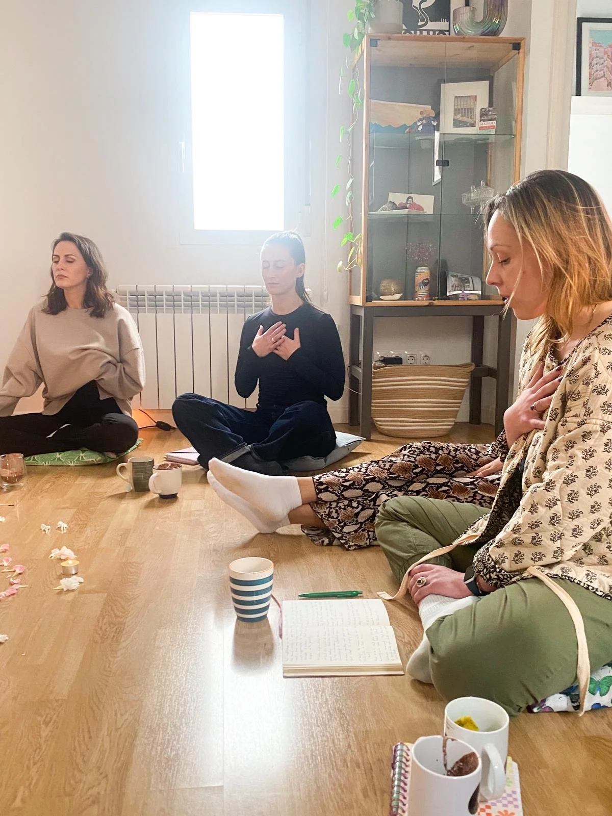 Three women sitting cross-legged on the floor with their hands over their hearts, participating in a meditation or mindfulness session. They are indoors with a large window, a wooden floor, and a cabinet in the background. Various cups and notebooks are placed on the floor around them.