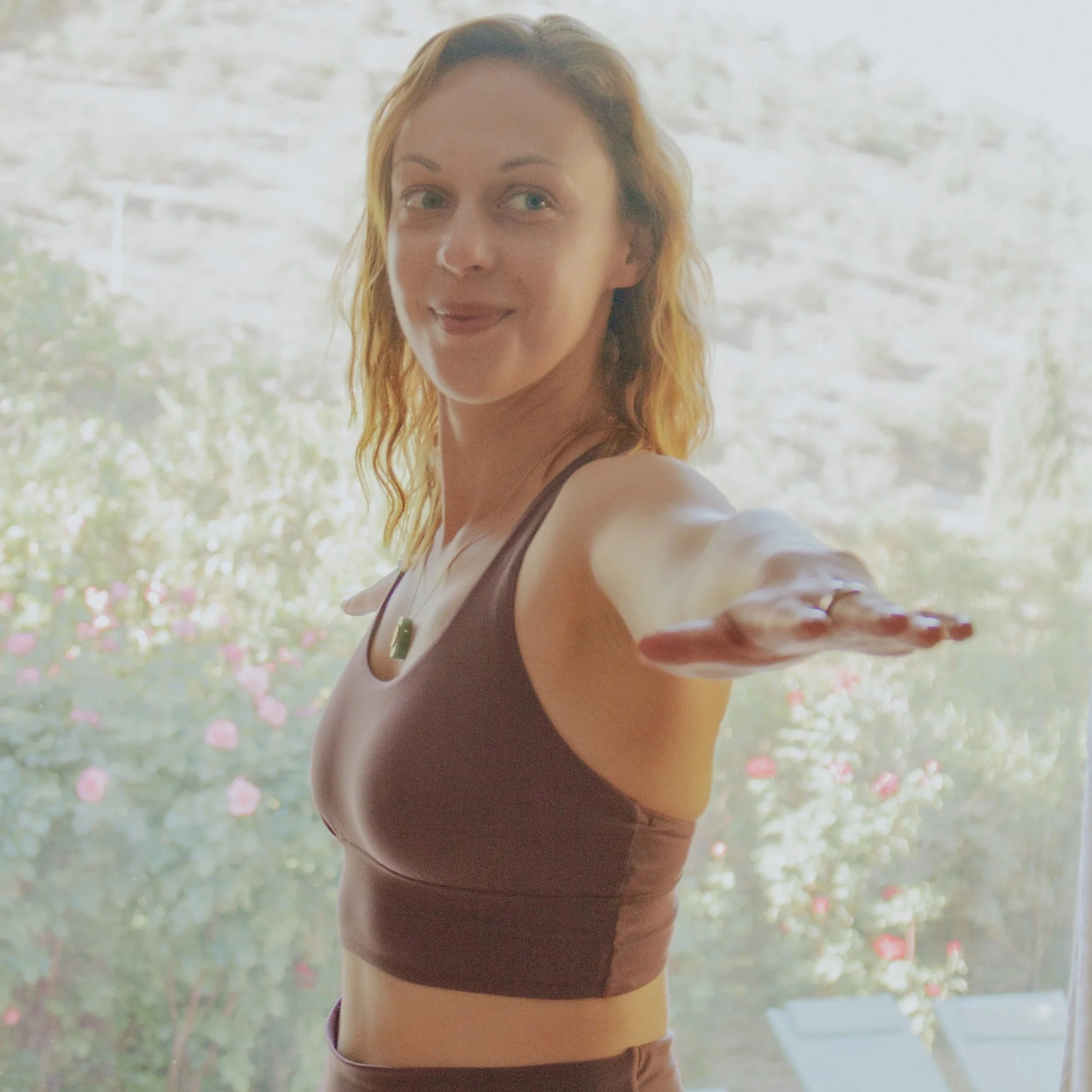 A woman with red hair in a brown athletic top stretching her arm outward indoors with a bright window in the background.