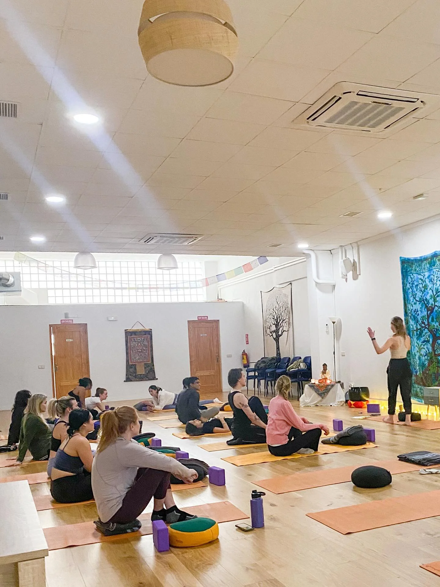 A group of people in a yoga class seated on yoga mats in a spacious room with wooden floors, listening to an instructor standing at the front near colorful wall hangings.