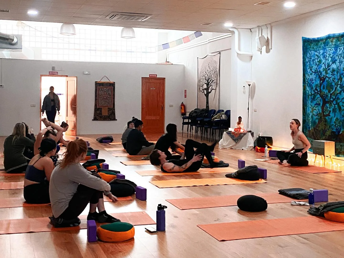 Group of people participating in a yoga class in a spacious room with wooden floors, colorful wall art, and relaxing lighting.