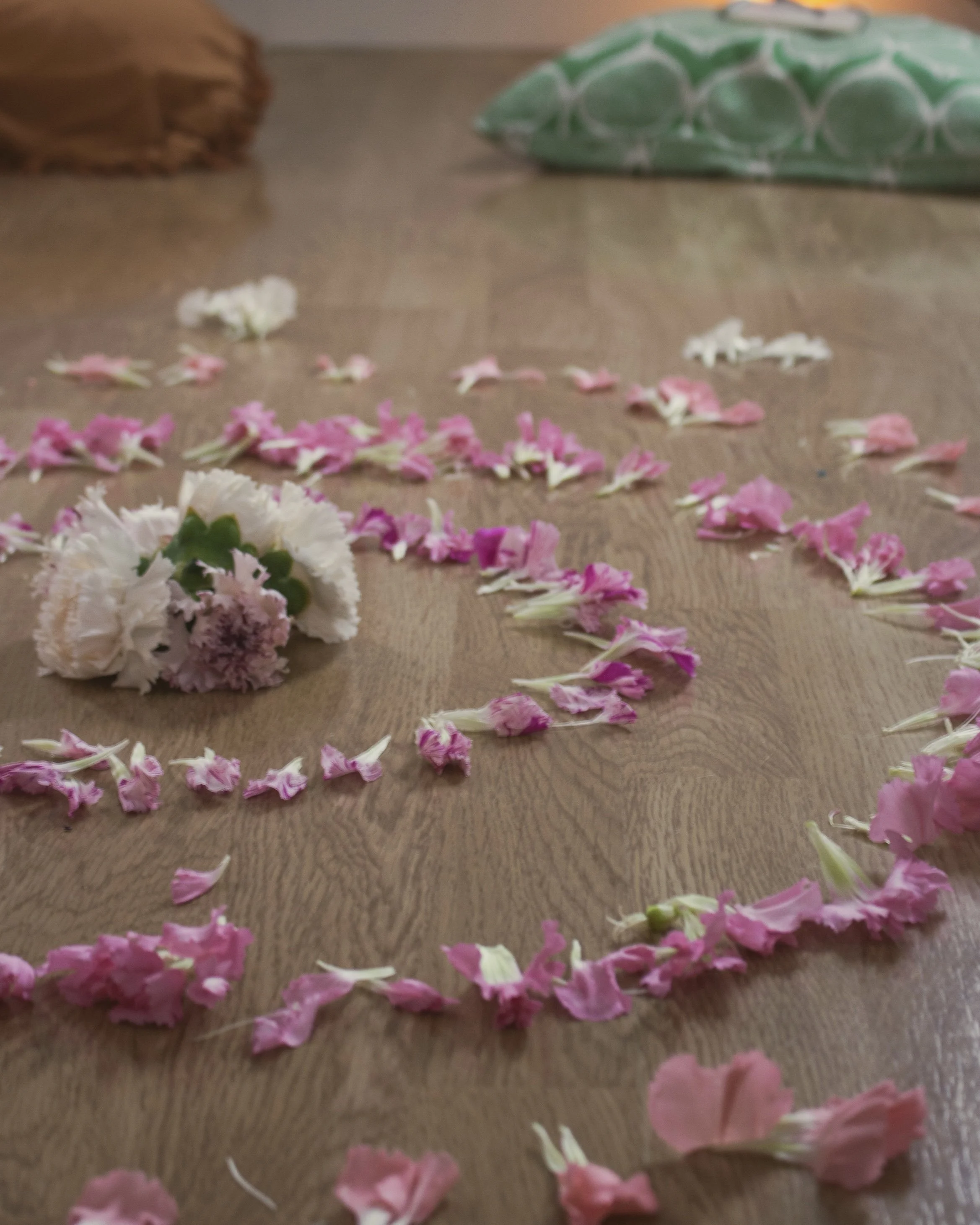 Pink and white flower petals arranged on a wooden floor, with a bouquet of white flowers in the foreground and a pillow in the background.