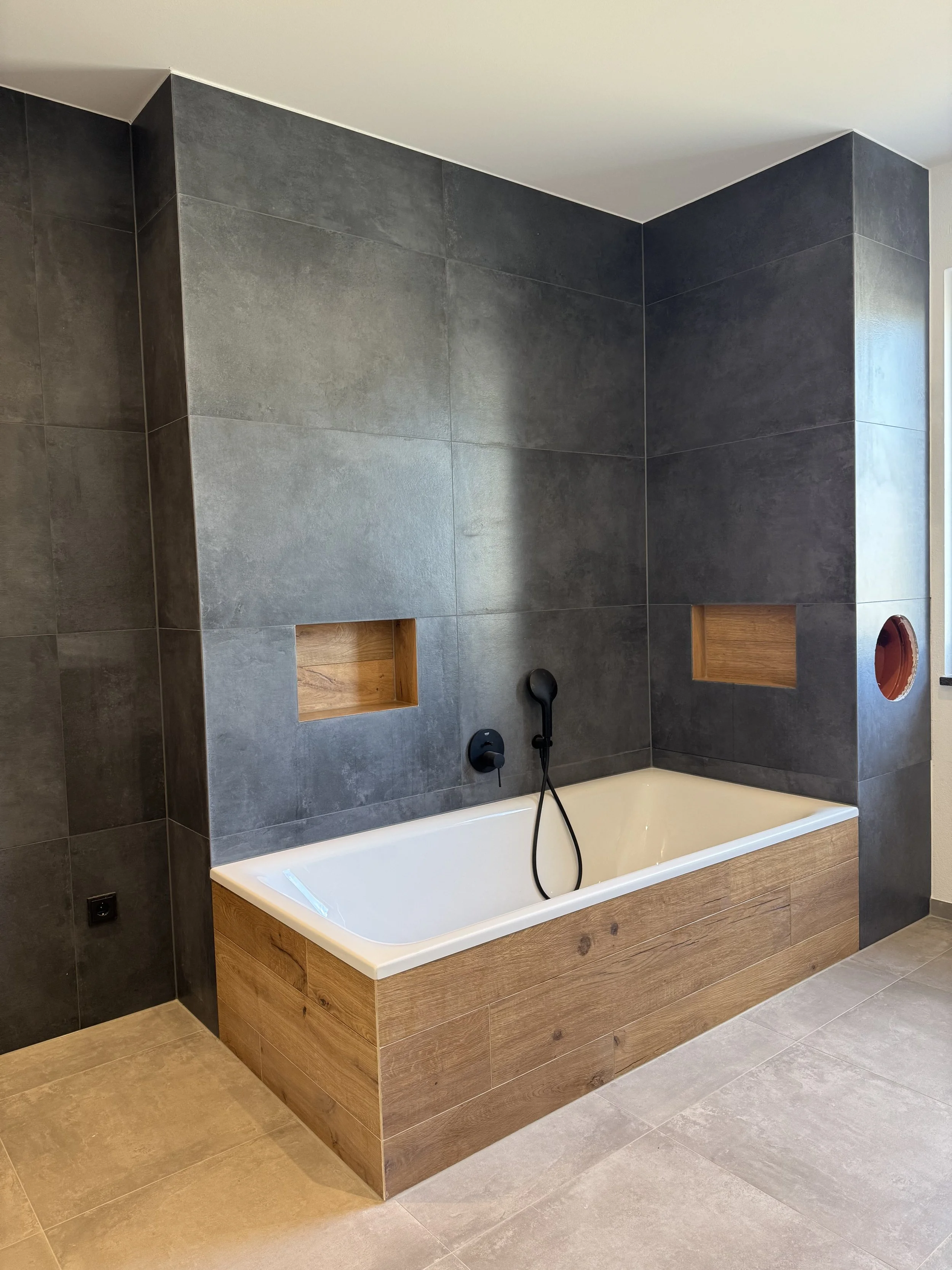 Modern Badezimmer mit dunklen Fliesen, Holzverkleidung um die Badewanne, zwei eingelassene Nischen in der Wand, Wasserhahn und Handbrause.