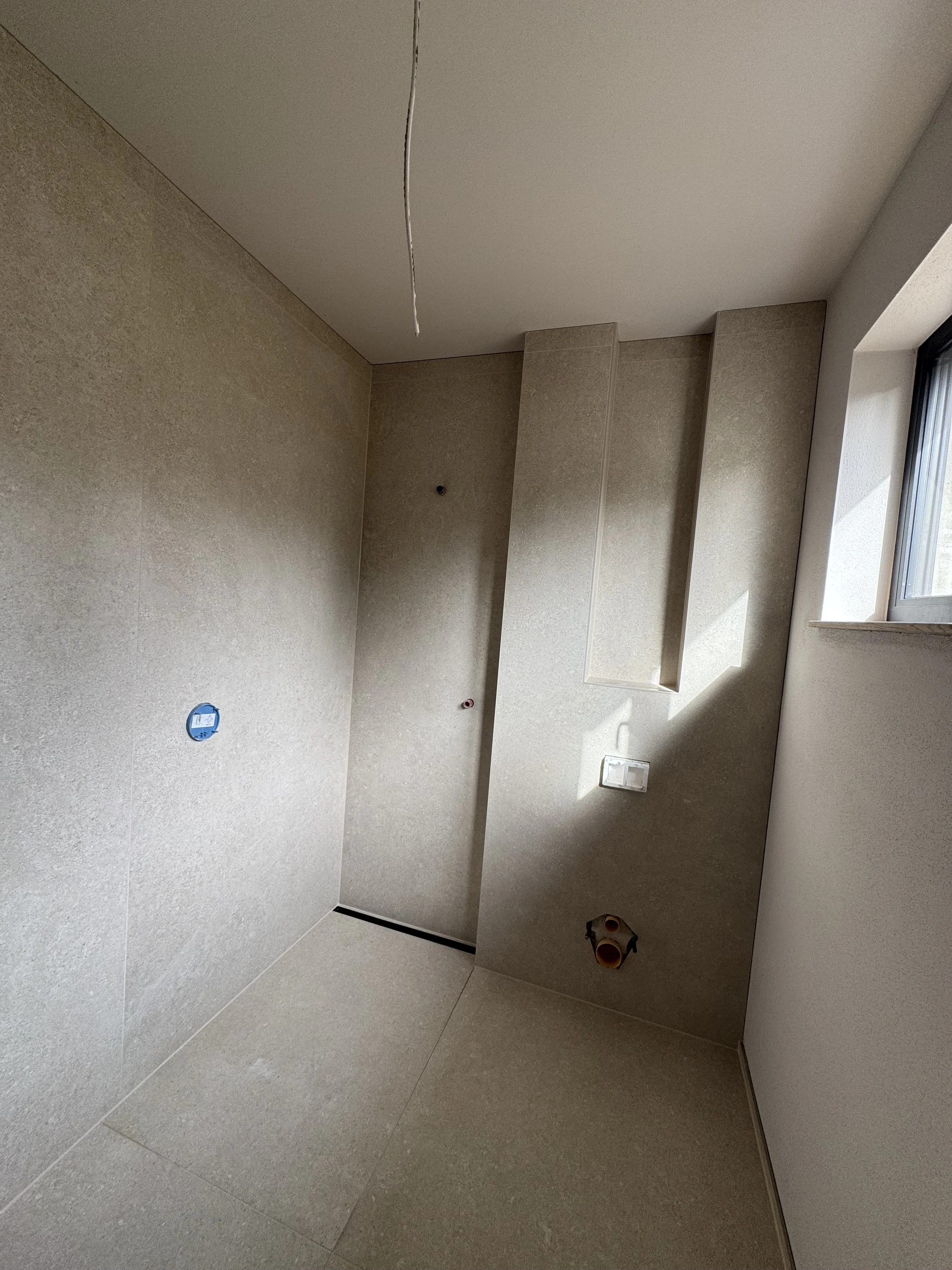 Innenraum eines fertiggestellten Badezimmers ohne Möbel, mit Wand- und Bodengestaltung, einer offenen Wasserleitung, einem Ausschnitt eines Fensters und einer Starkstromdose.