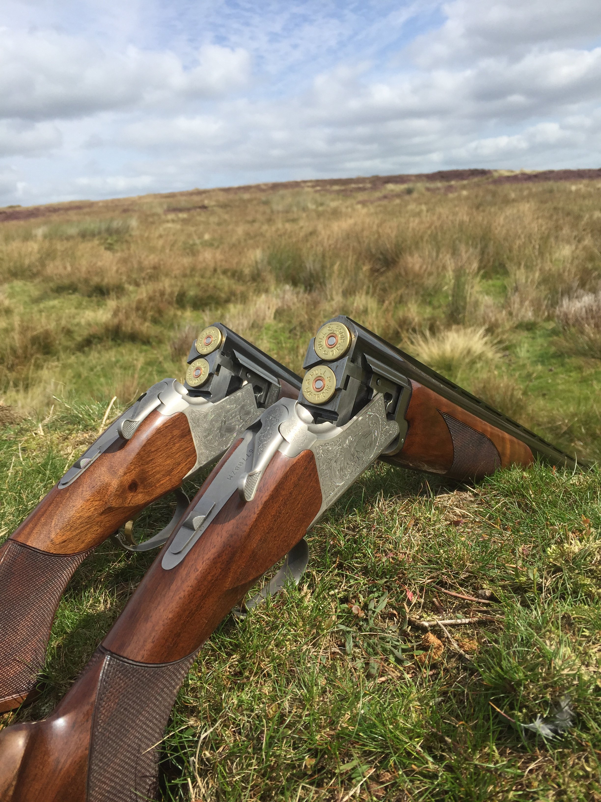 Two handguns with wooden grips and silver decorative engravings, lying on grass in an open field with cloudy sky.
