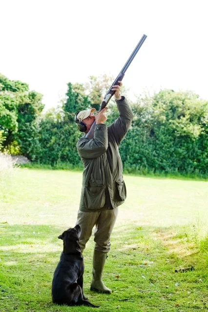 A person wearing ear protection, glasses, and outdoor clothing is aiming a shotgun into the sky in an open grassy area, with a black dog sitting nearby.