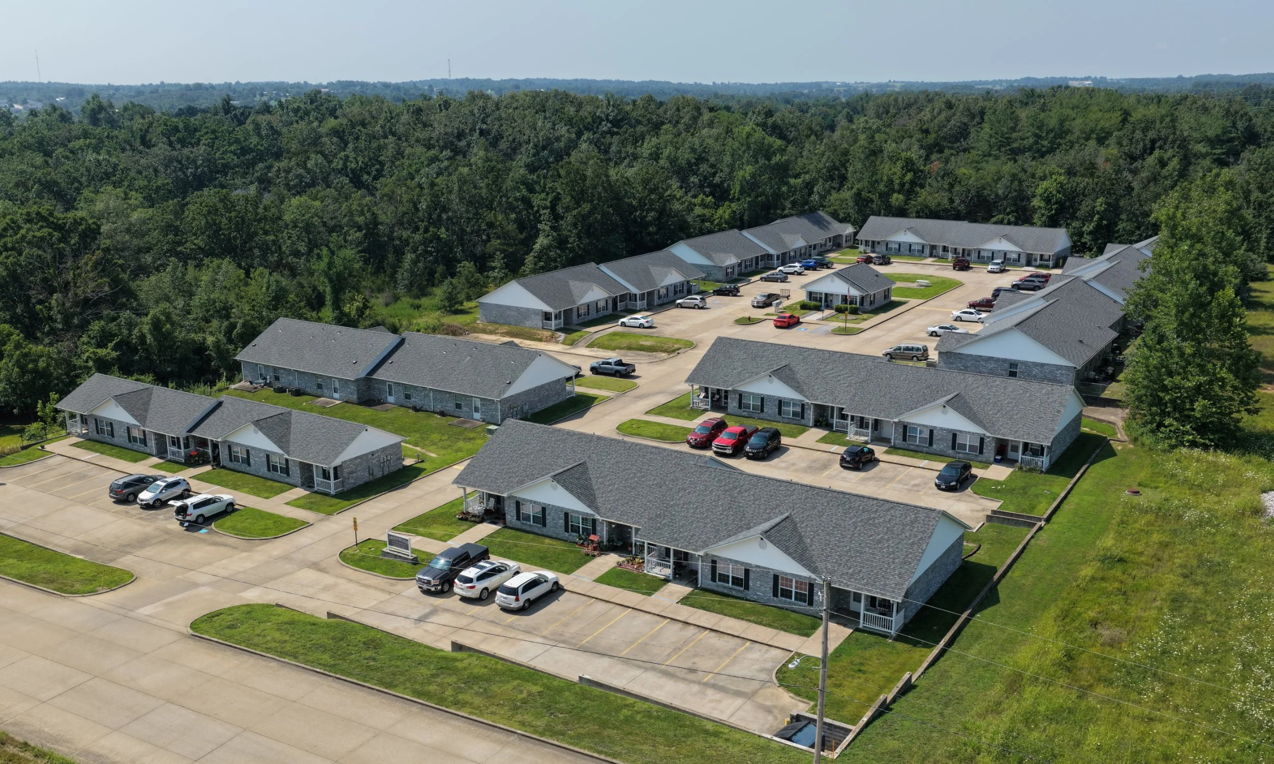 Aerial view of a residential complex with multiple single-story buildings, parking lots, and surrounding trees.