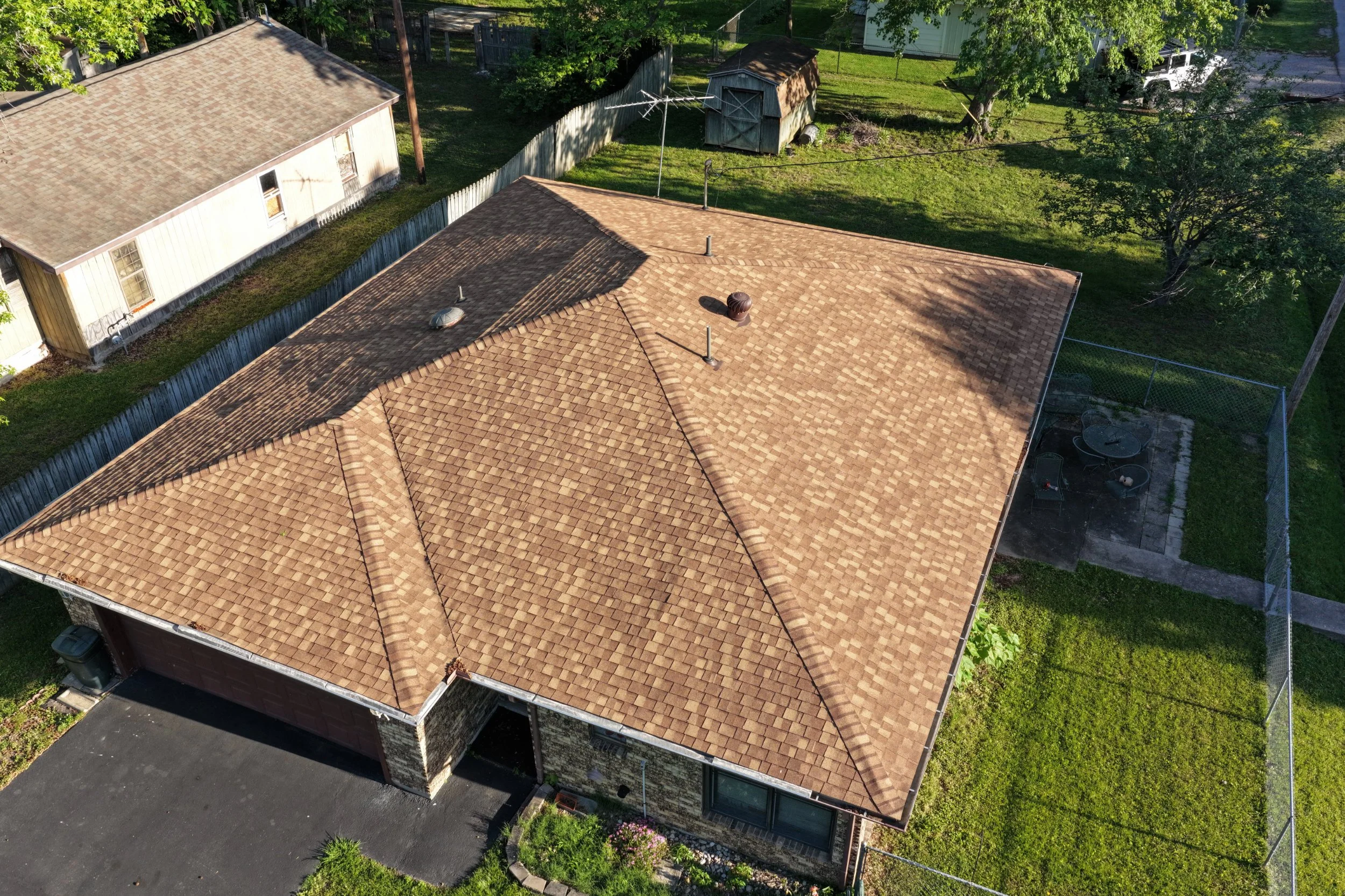 Aerial view of a house with a brown tiled roof, surrounded by green trees and a backyard with a shed and a fenced area.