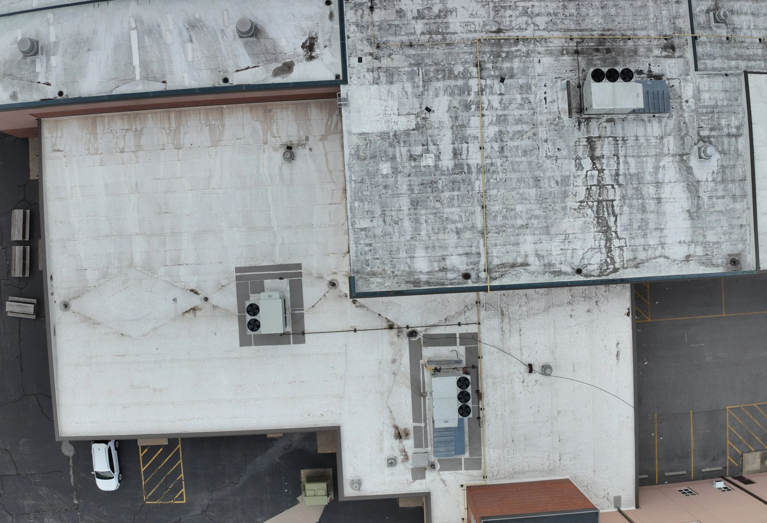 Aerial view of a building's rooftop with weathered white surface, various HVAC units, vents, yellow communication cables, and a parked white vehicle in the parking lot below.