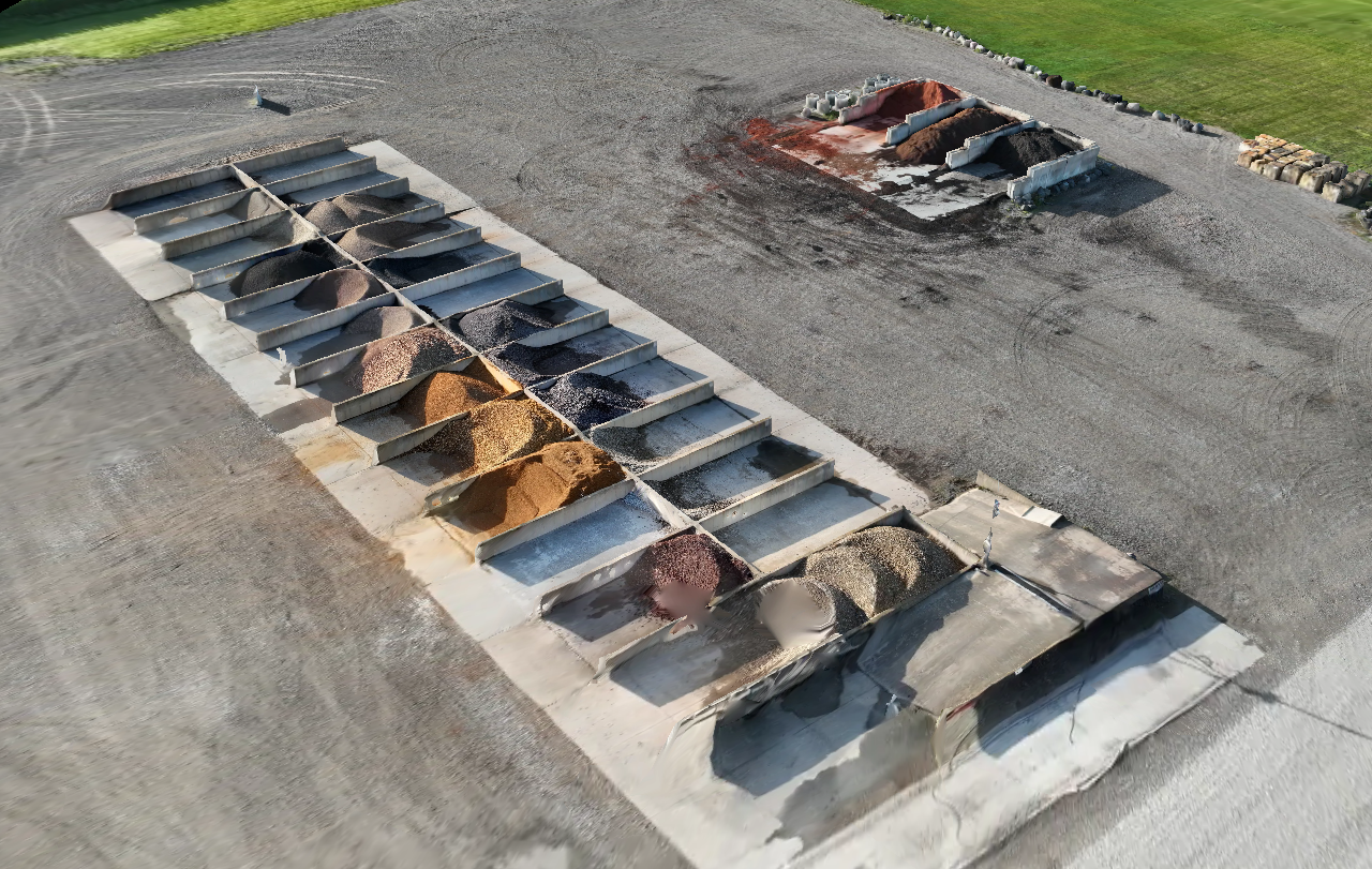 Construction site with multiple piles of different colored gravel and dirt organized in concrete bins, and a raised area with loose soil or gravel in background.