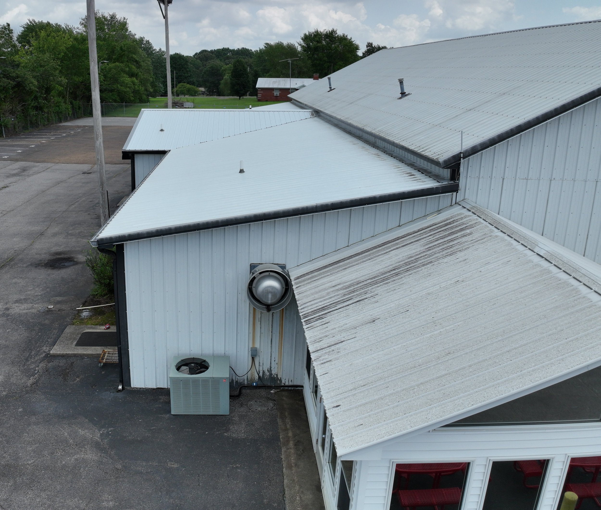 Aerial view of a building with white metal roofs and visible vents, located near a parking lot and surrounded by trees and open green space.
