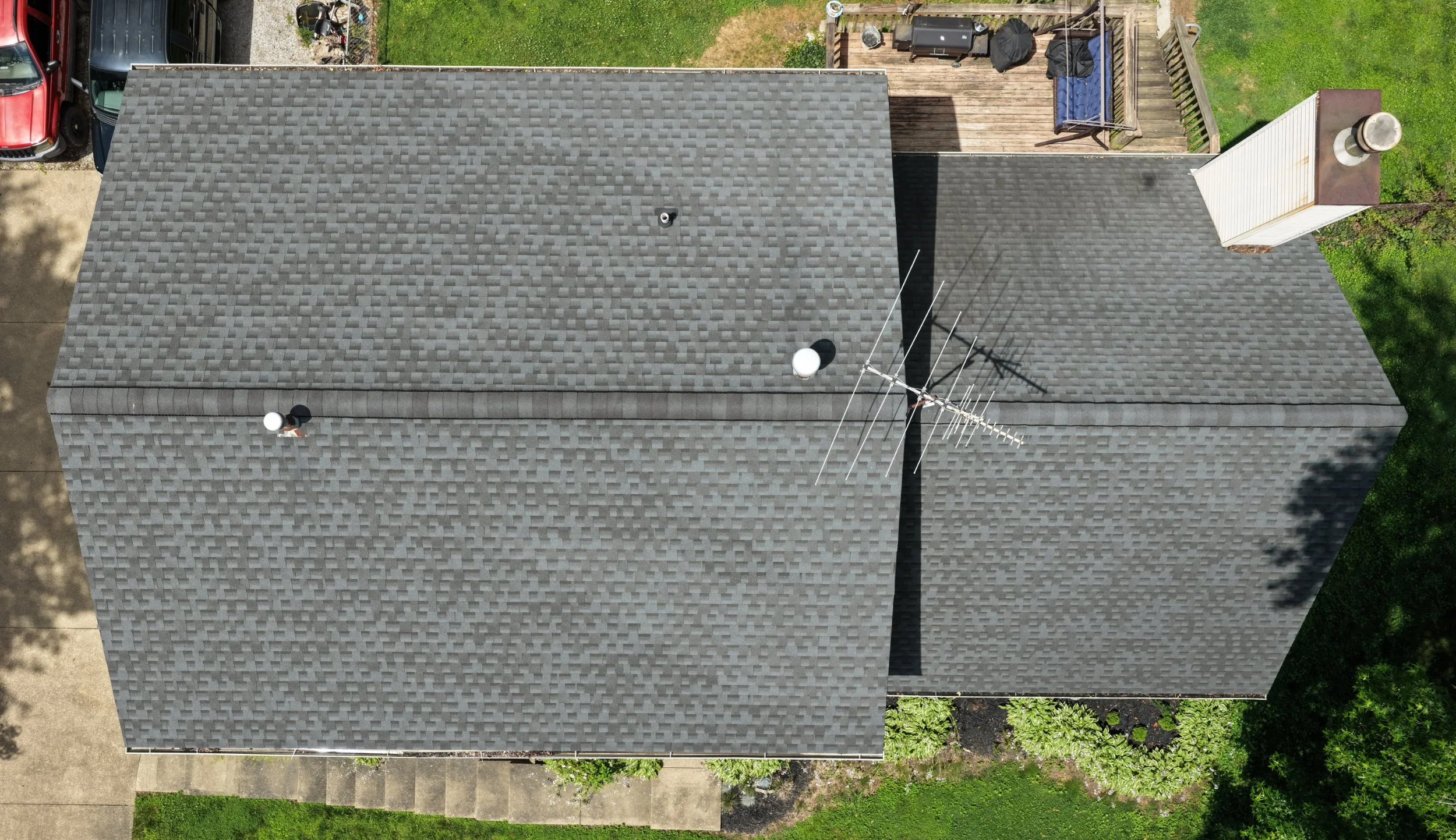 An aerial view of a house roof with gray shingles, two roof vents, and satellite dish, surrounded by green grass, driveway, and a backyard with a deck, barbecue grill, and outdoor furniture.