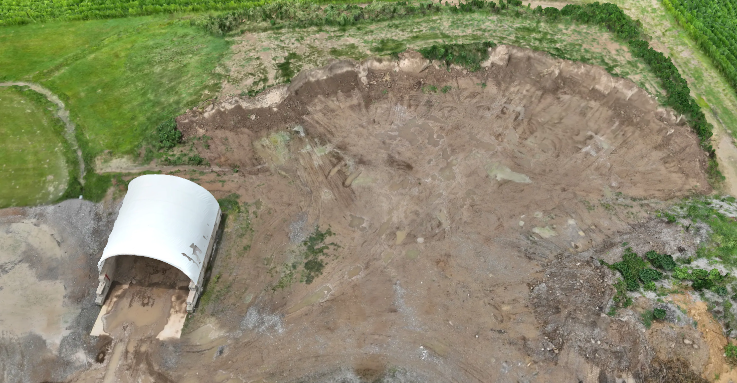 An aerial view of a construction site with a large, partially excavated area and a white excavator shelter nearby, surrounded by green fields and plants.
