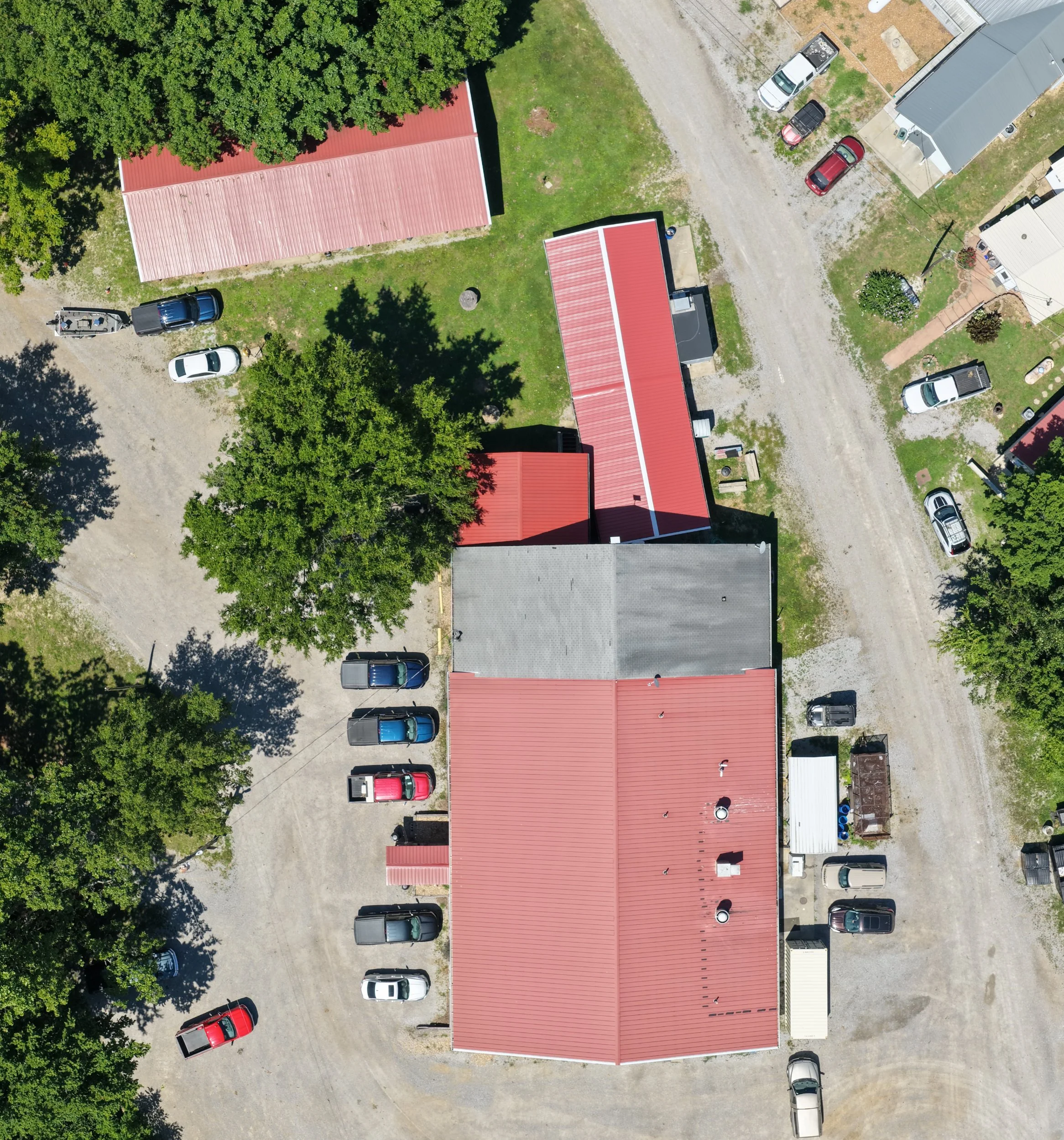 An aerial view of a residential area showing multiple vehicles parked along a dirt road, several houses with red roofs, green trees, and patches of grass.