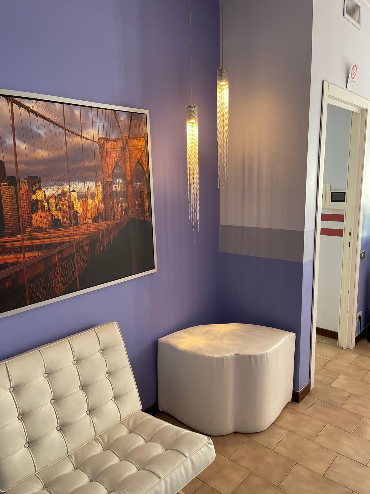 Sala d'attesa con pareti viola, quadro di New York, due lampadari con cristalli pendenti, divano bianco e una sedia bianca.