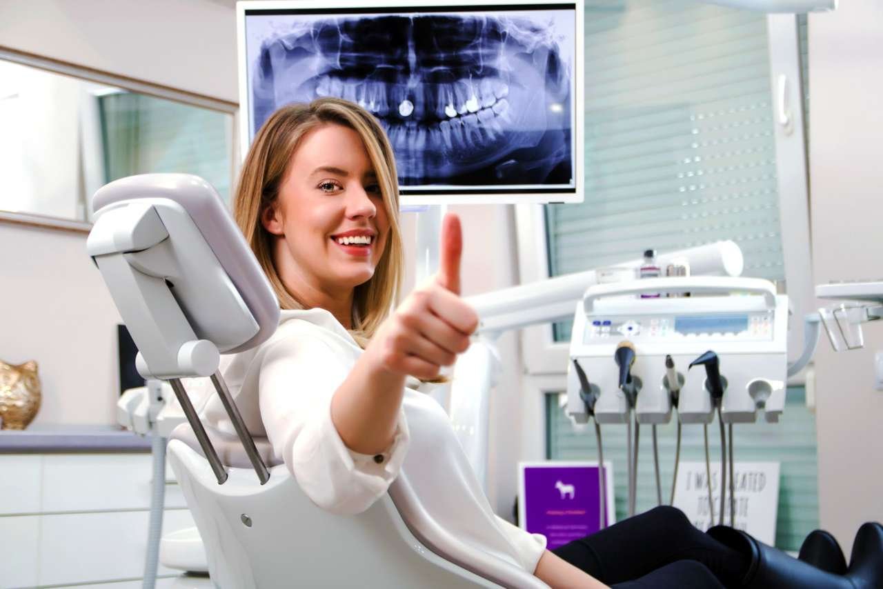 Donna sorridente in uno studio dentistico, che fa il gesto del pollice in su, con strumentazione dentistica e radiografia dentale sullo sfondo.