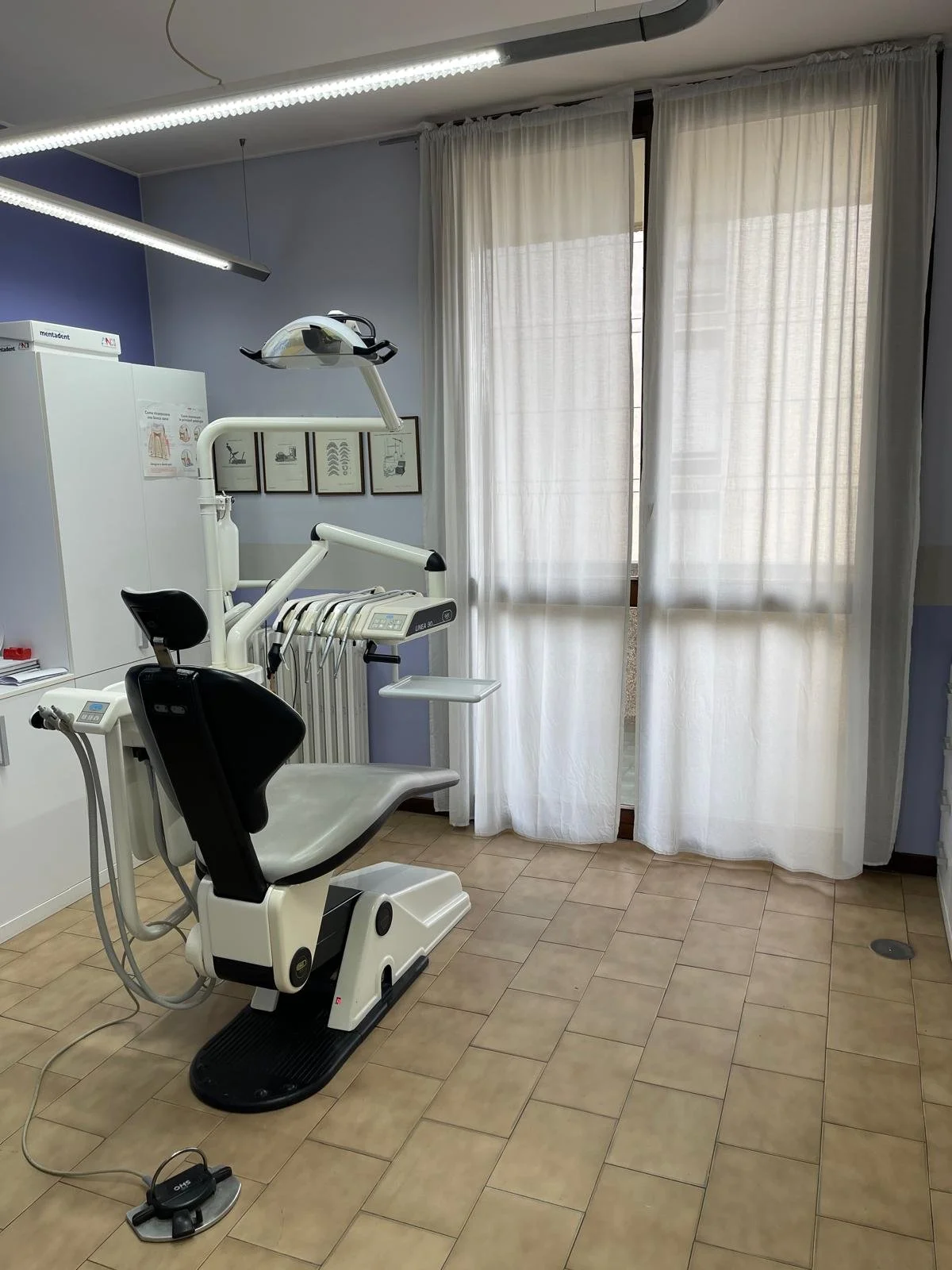 Ambiente odontoiatrico con sedia dentistica e attrezzature, luci e finestre con tende bianche.