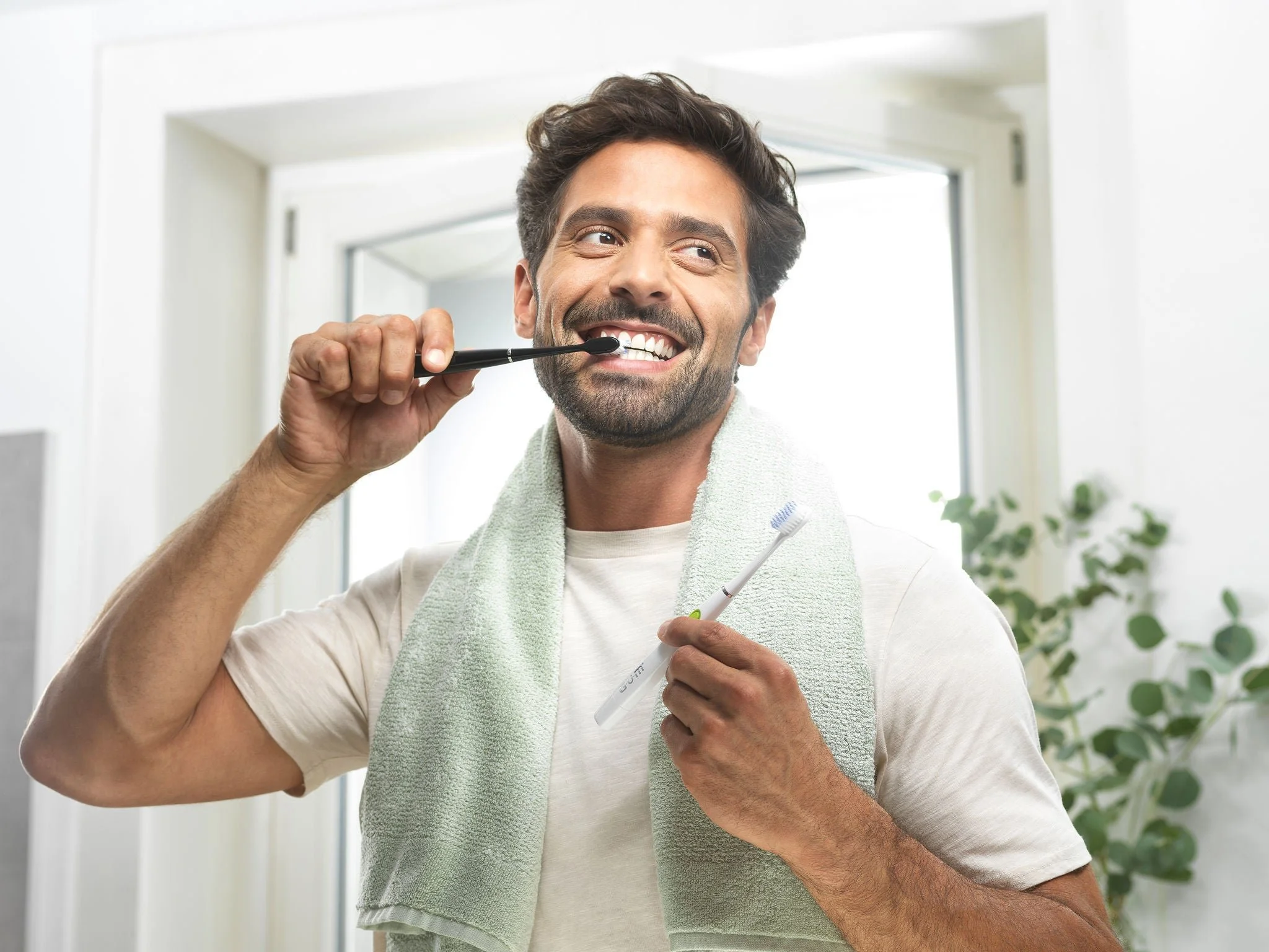 Uomo che si spazzola i denti con una spazzola elettrica, tenendo anche un vecchio spazzolino in mano, con una salvietta intorno al collo, in un bagno luminoso.