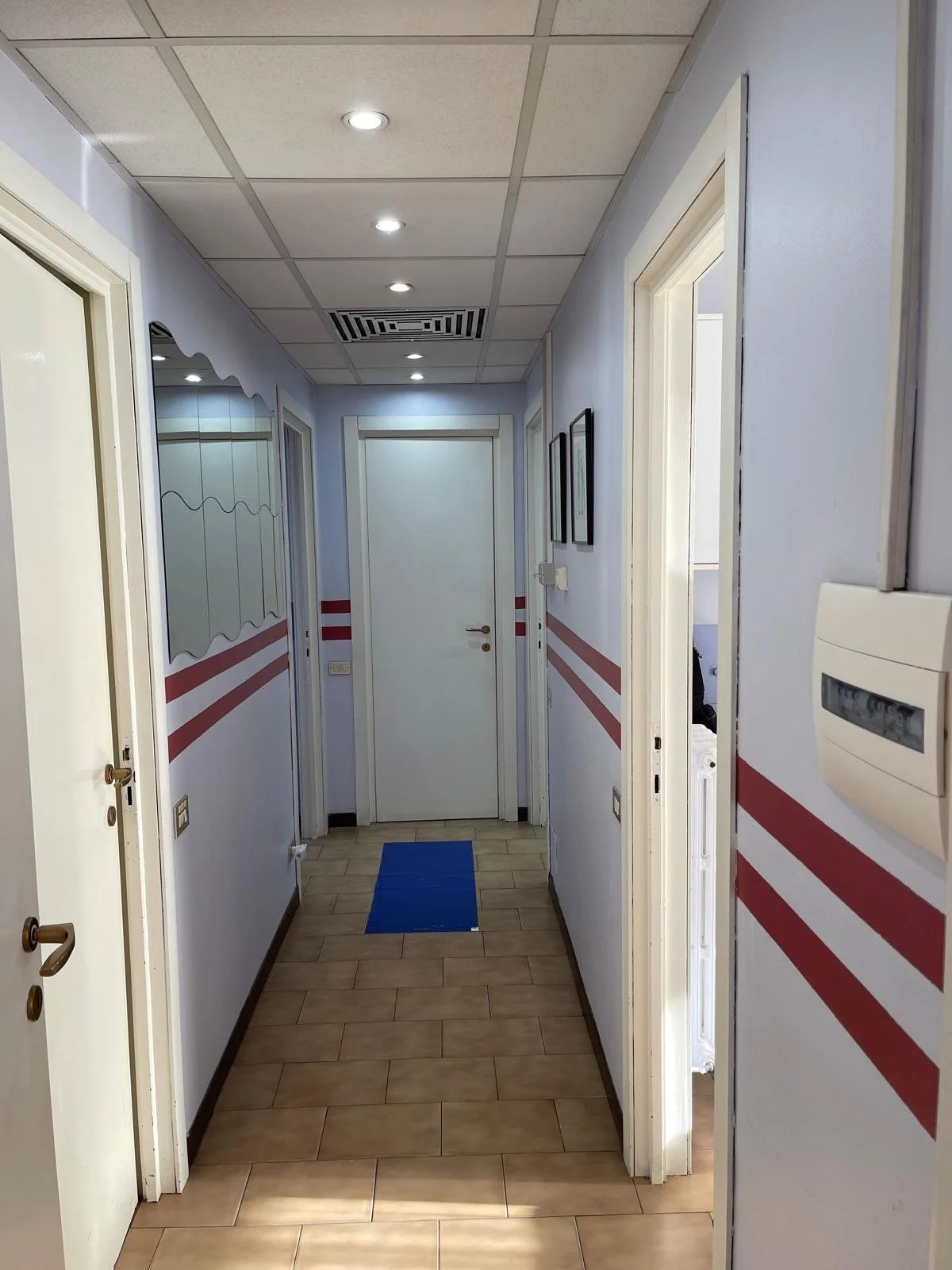 Corridoio interno di un edificio con pareti azzurre, una mobilia a specchio sulla sinistra e tre quadri neri appesi sulla destra. Il soffitto è composto da pannelli con luci e una ventilazione. La pavimentazione è in piastrelle marroni e c'è un tappe