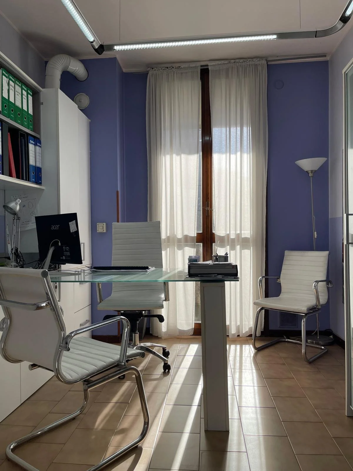 Ufficio con scrivania in vetro, sedia girevole, due sedie bianche con braccioli, mensole con cartelle, computer e lampade, pareti azzurre e finestra con tende bianche.